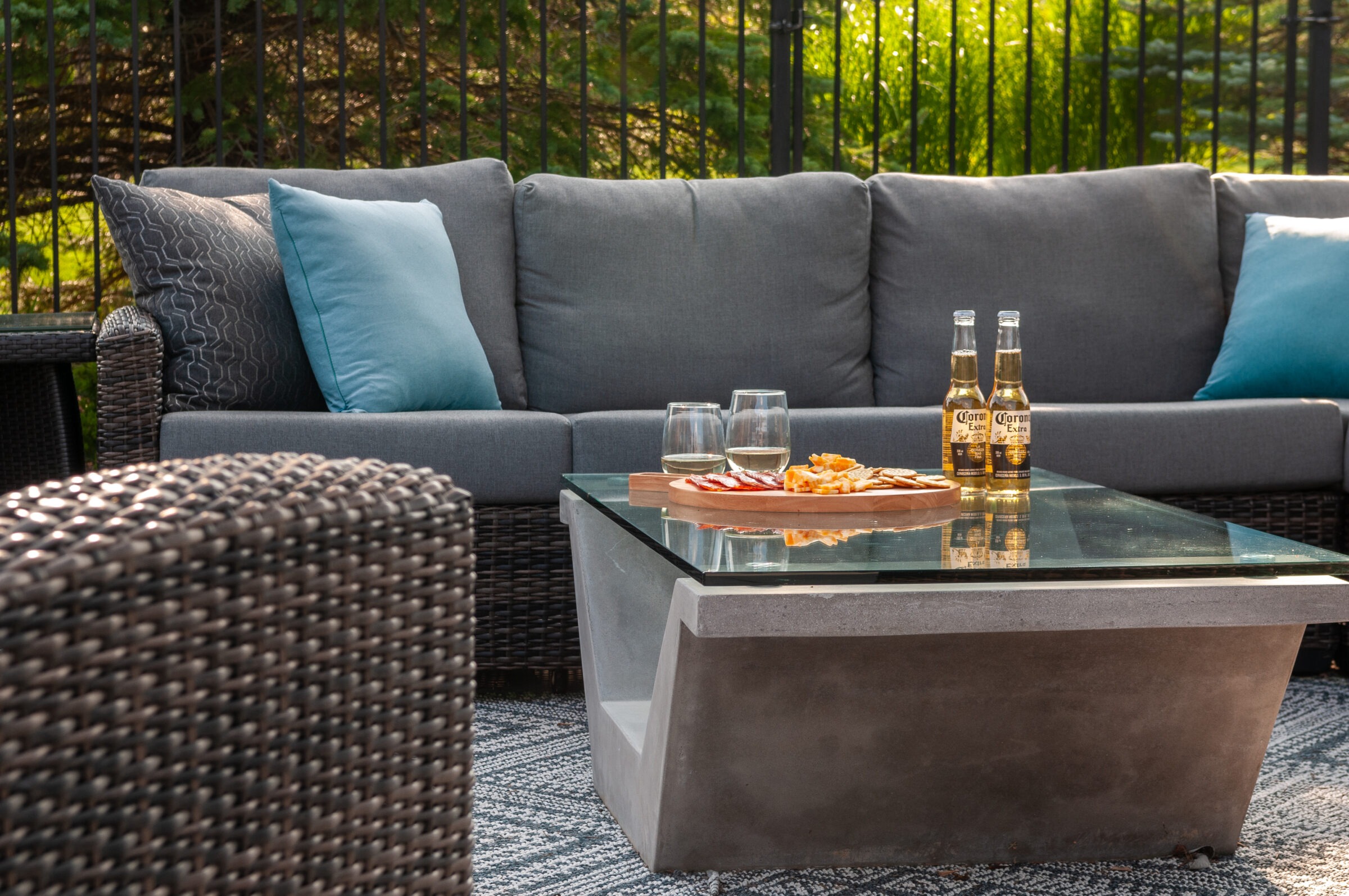 Outdoor patio scene with wicker furniture, glass and concrete table, snacks, drinks, blue cushions, and greenery in the background. Relaxed and inviting atmosphere.