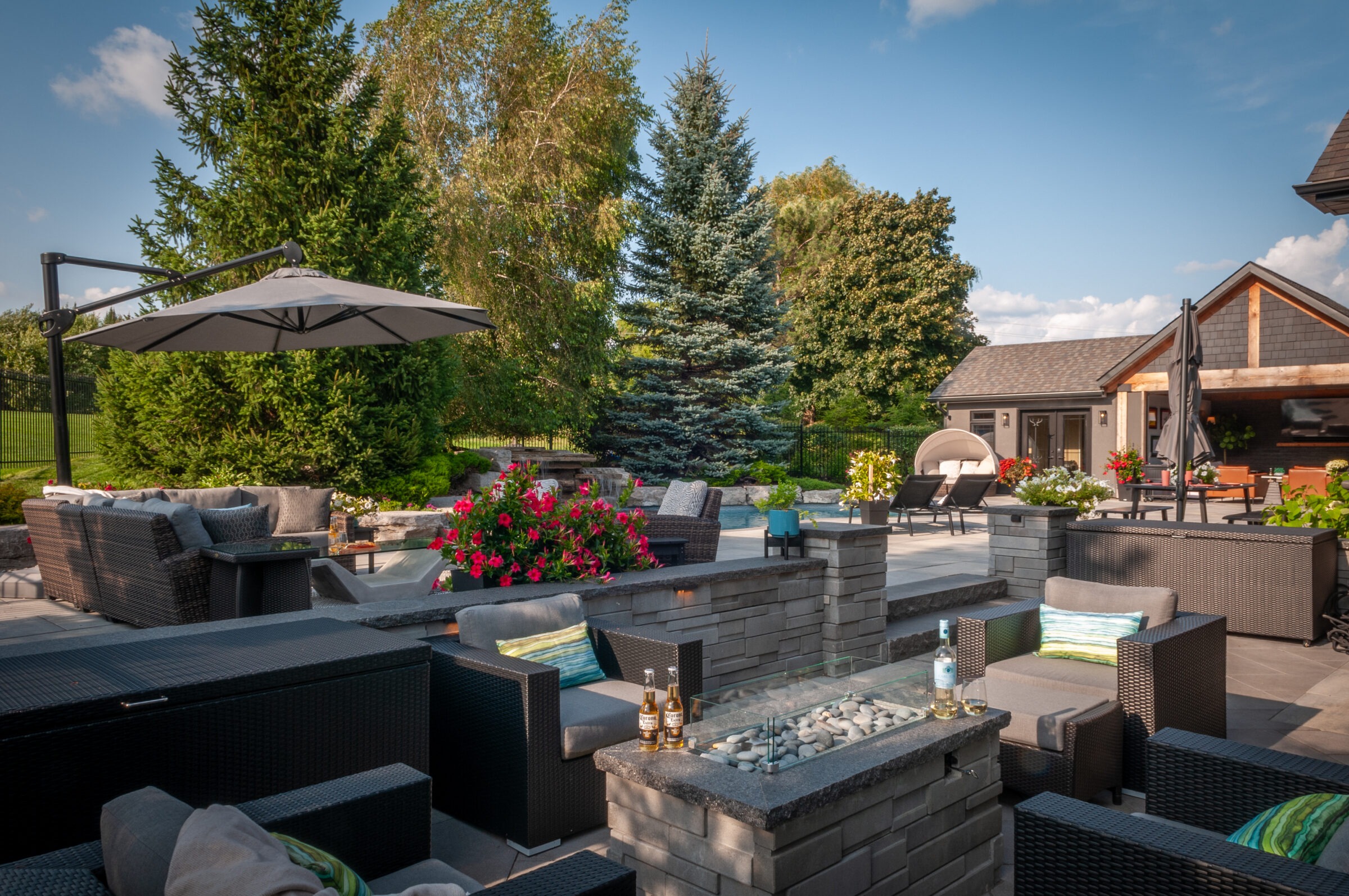 Luxurious backyard patio with modern furniture, fire pit, umbrellas, and vibrant flowers, surrounded by lush greenery and a small wooden structure.