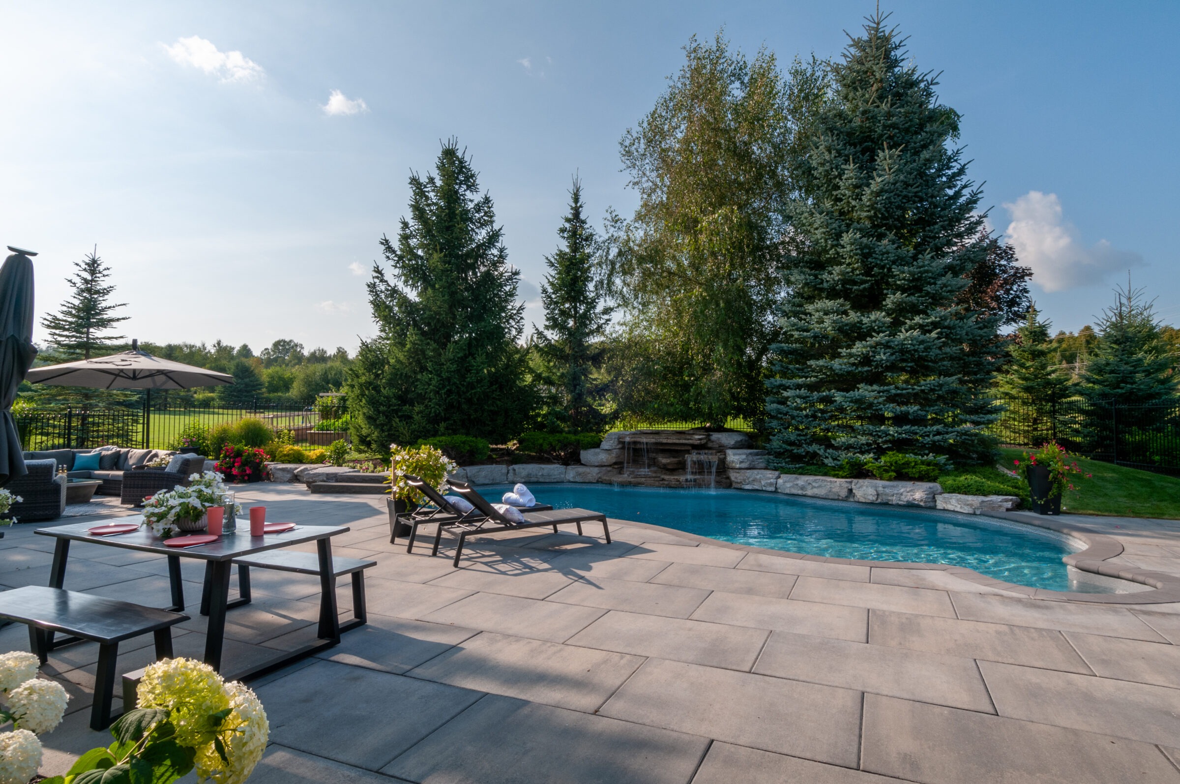 A serene backyard features a swimming pool, lounge chairs, dining area, and lush greenery under a clear blue sky.