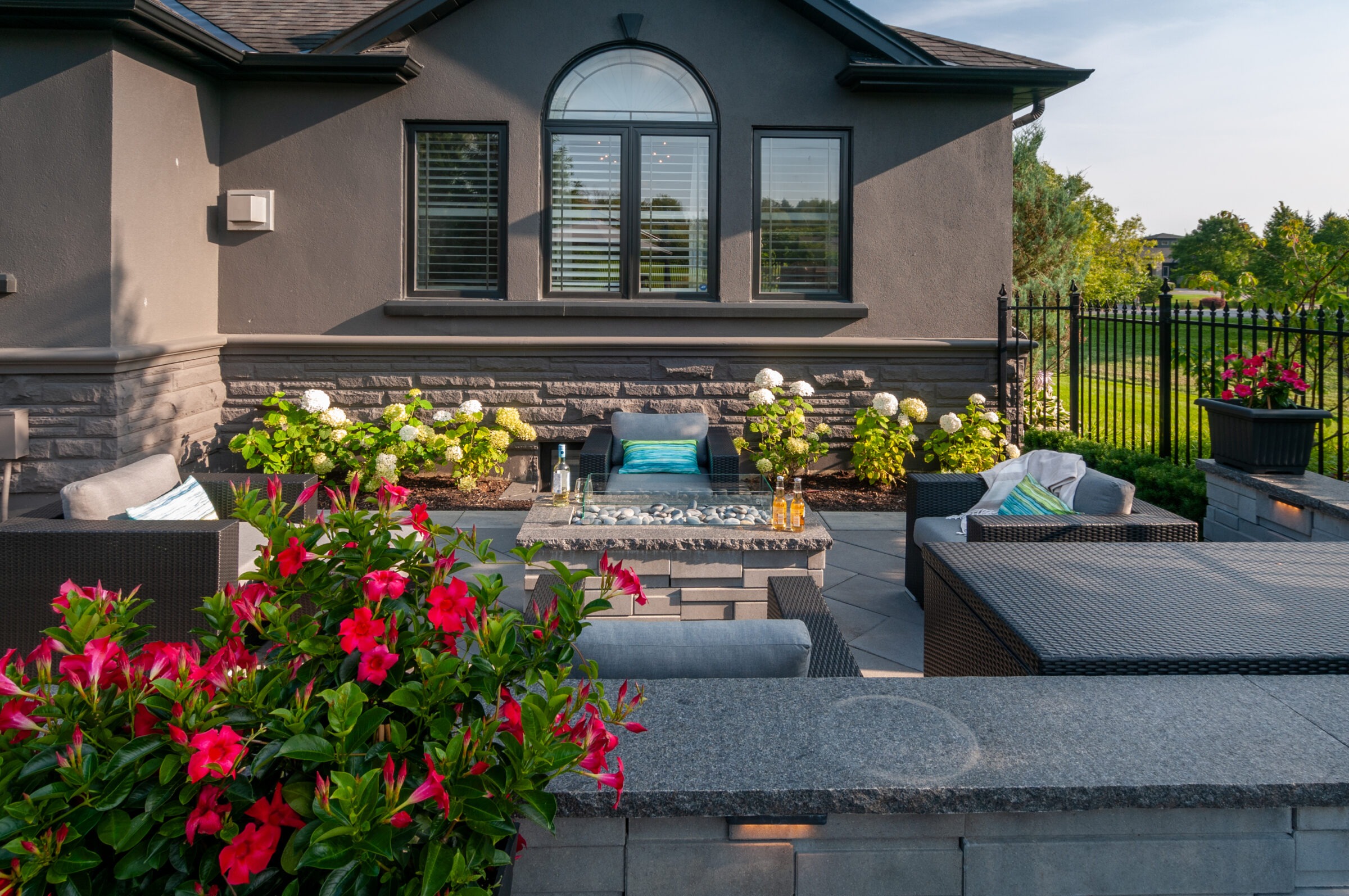 A modern backyard patio with grey seating, vibrant flowers, and a rectangular stone fire pit, surrounded by lush greenery and iron fencing.
