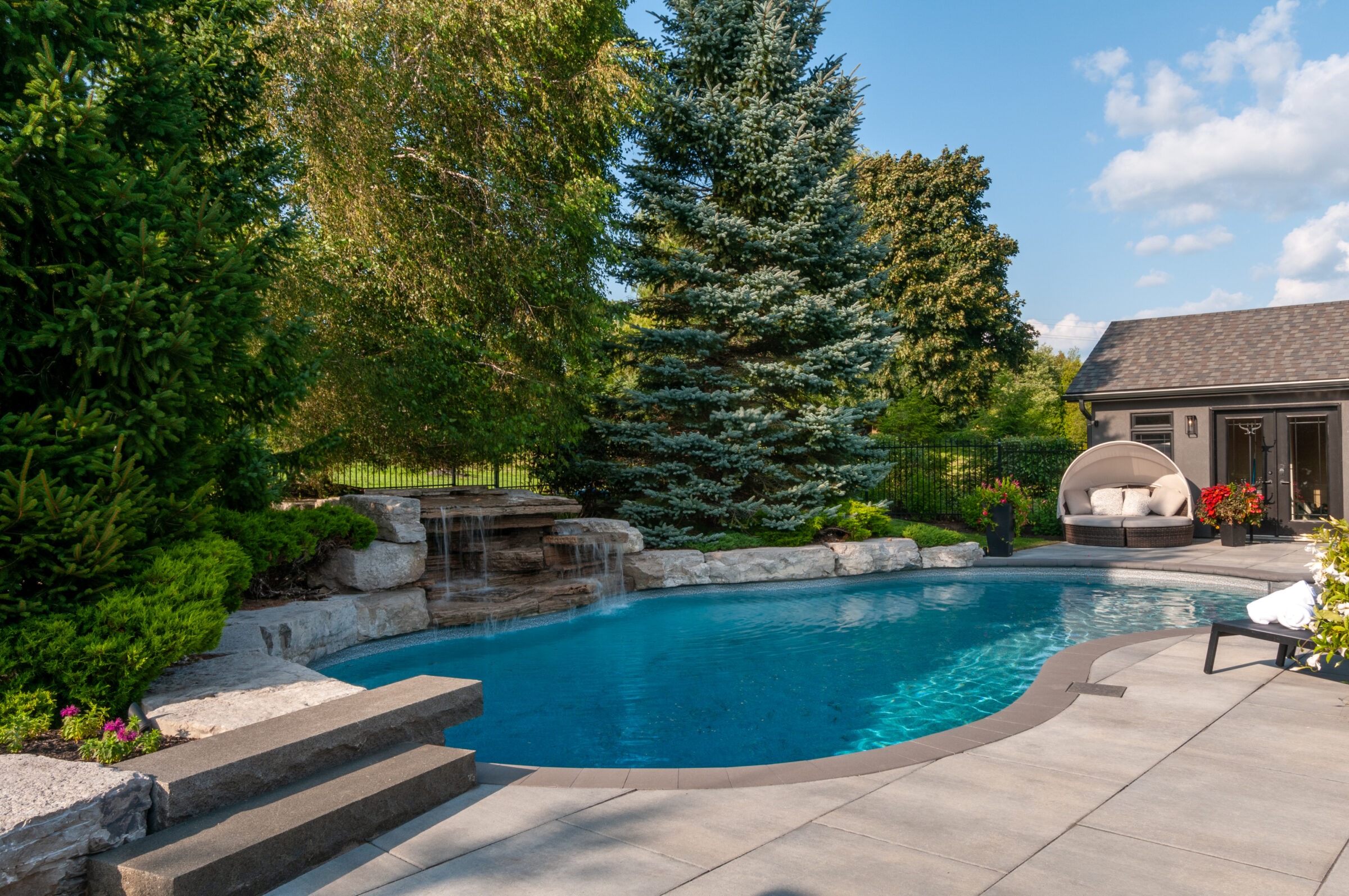 A serene backyard pool with a waterfall, surrounded by lush trees, a cozy lounge area, and a small pool house.