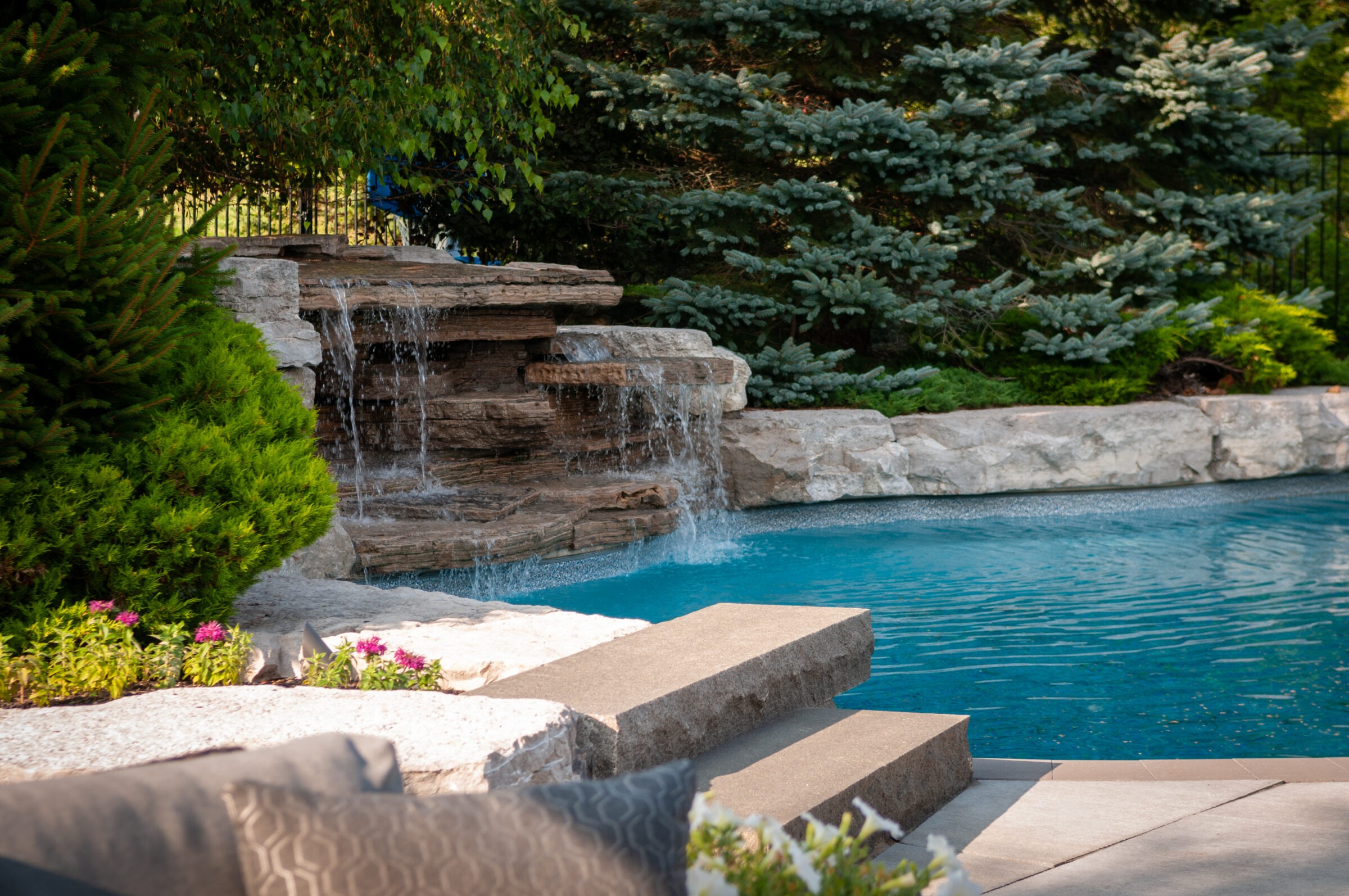 A serene backyard features a cascading rock waterfall, vibrant green foliage, steps leading into a clear blue pool, and colorful flowers.