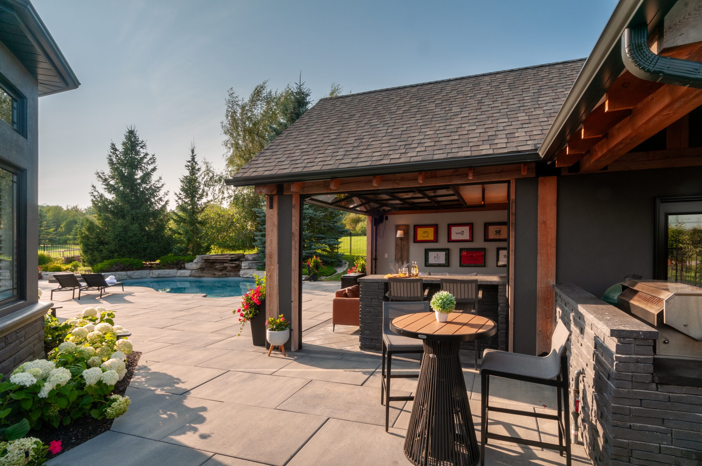 Modern patio with a pool, surrounded by trees and flowers. Lounge chairs and outdoor bar under a wooden roof create a relaxing atmosphere.