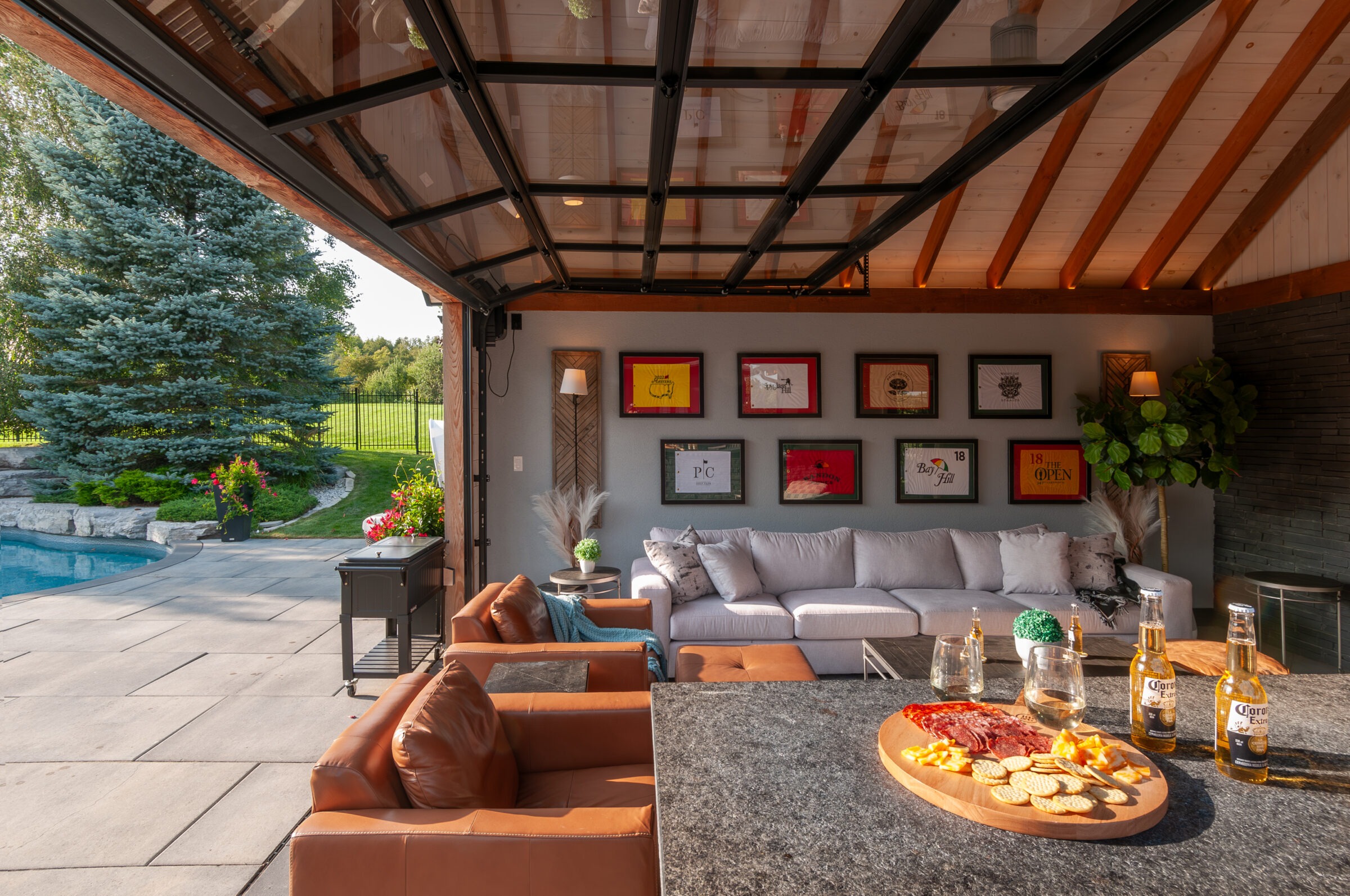 Stylish backyard patio with seating, pool, and snacks on the table. Decorated walls; sunny garden view from open garage-style door.
