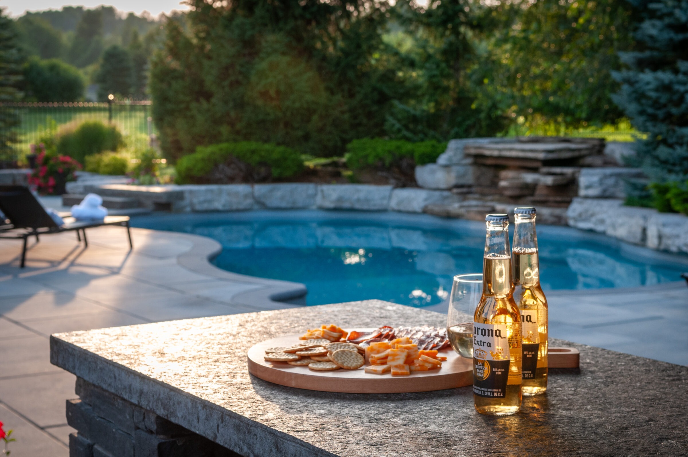 A serene backyard pool scene with a table featuring cheese, crackers, and beverages, surrounded by lush greenery and lounge chairs.