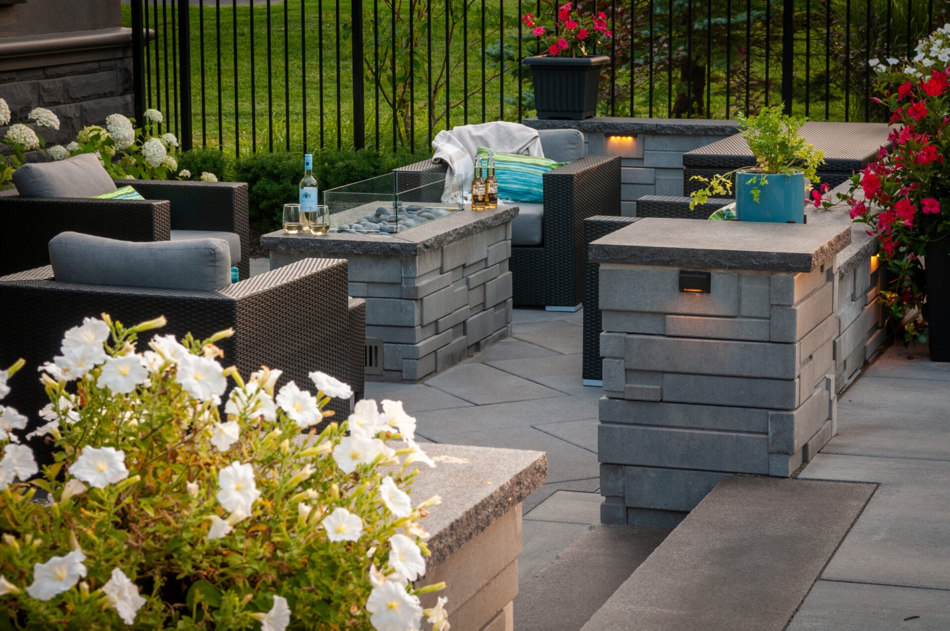 Outdoor patio with wicker furniture, cushions, and a glass fireplace. Surrounded by vibrant flowers and potted plants, creating a cozy ambience.