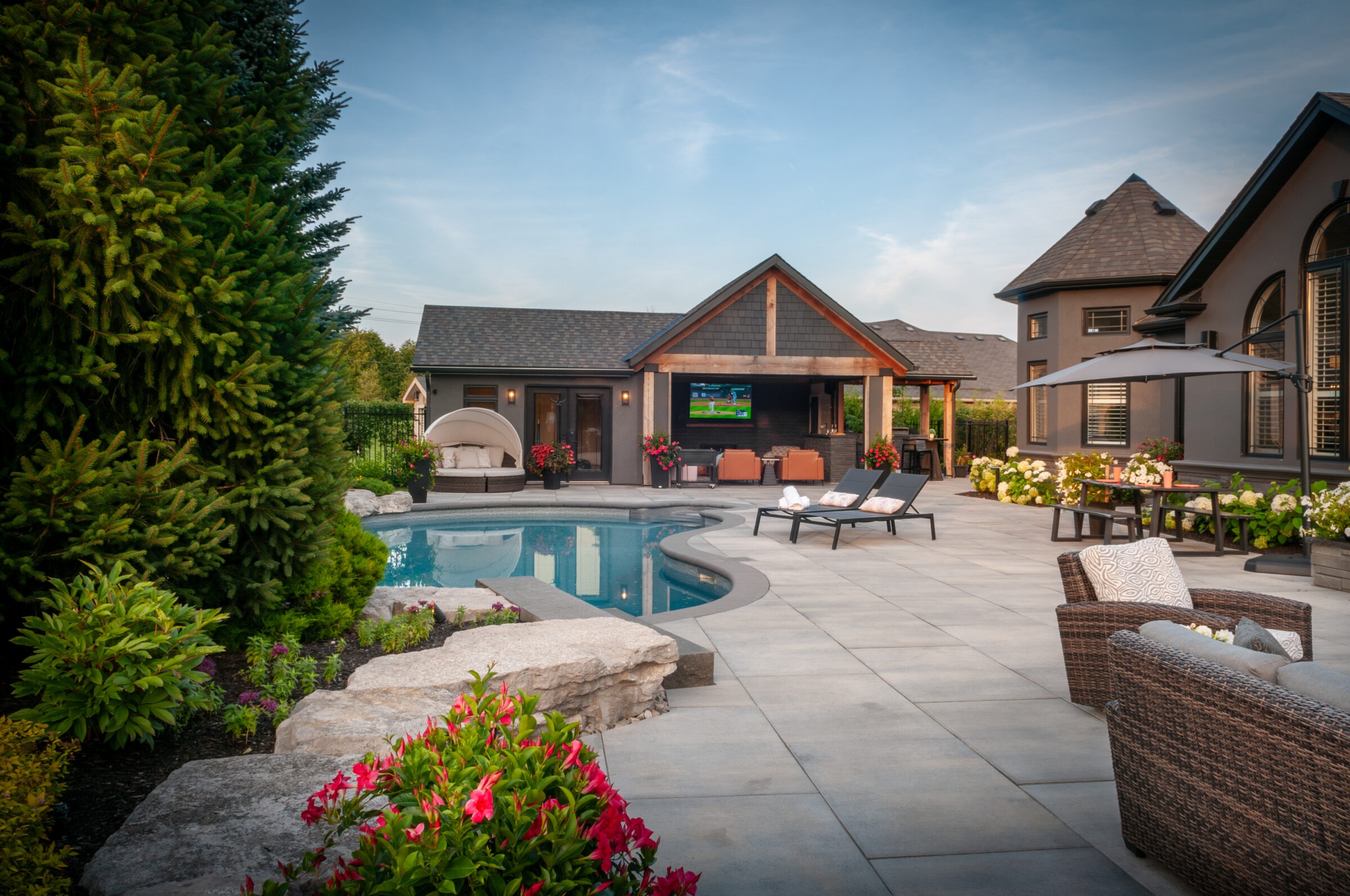 Luxurious backyard with a swimming pool, lounge chairs, and a cabana. Beautiful landscaping includes trees and vibrant flowers. Modern outdoor living space.