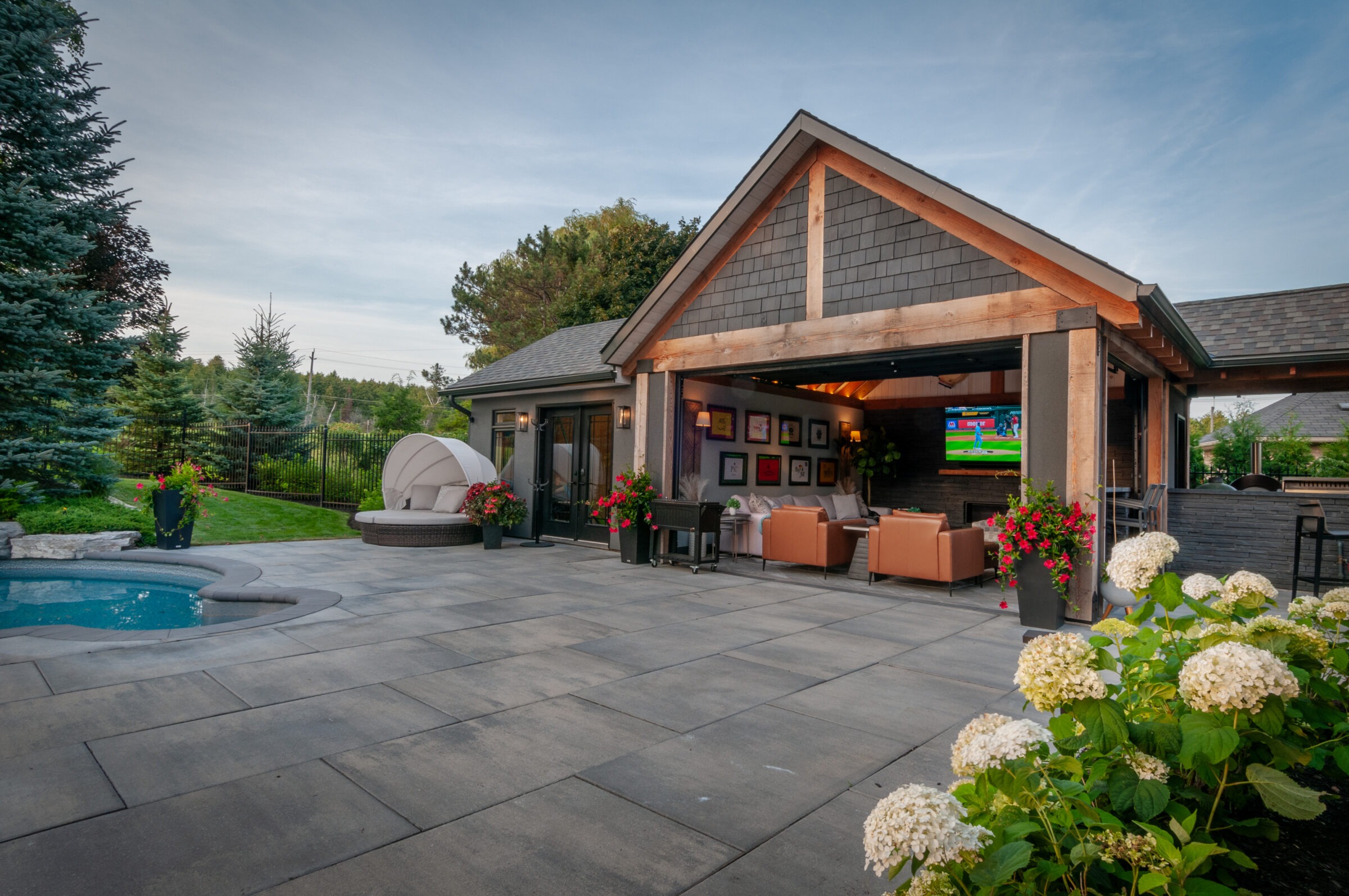 A cozy backyard patio with seating, a pool, greenery, and a TV under a wooden pavilion. Flowers enhance the tranquil atmosphere.
