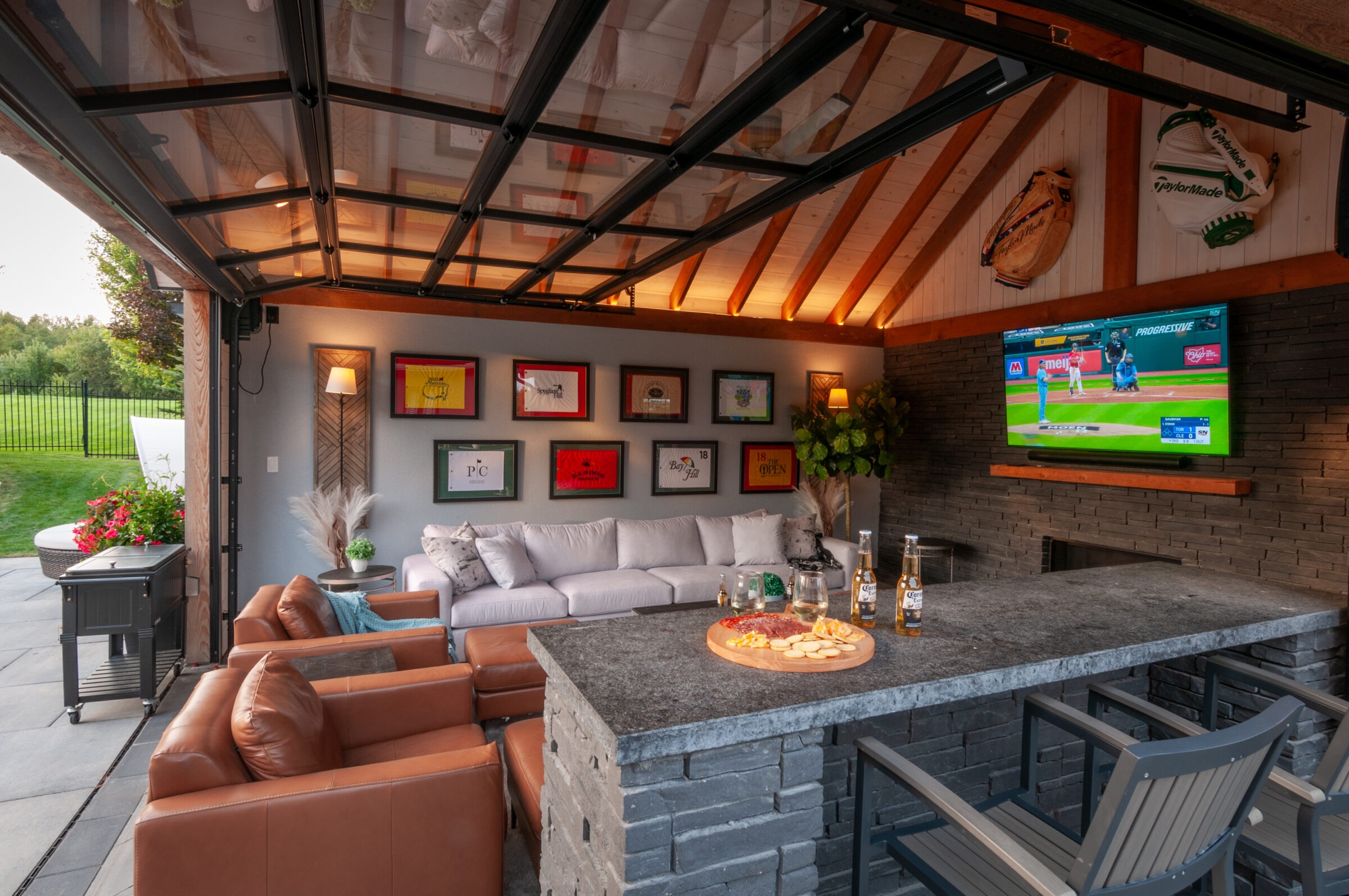 Cozy, open-air living space with leather chairs, TV displaying baseball, artwork on walls, a spacious sofa, and a stone bar with snacks.