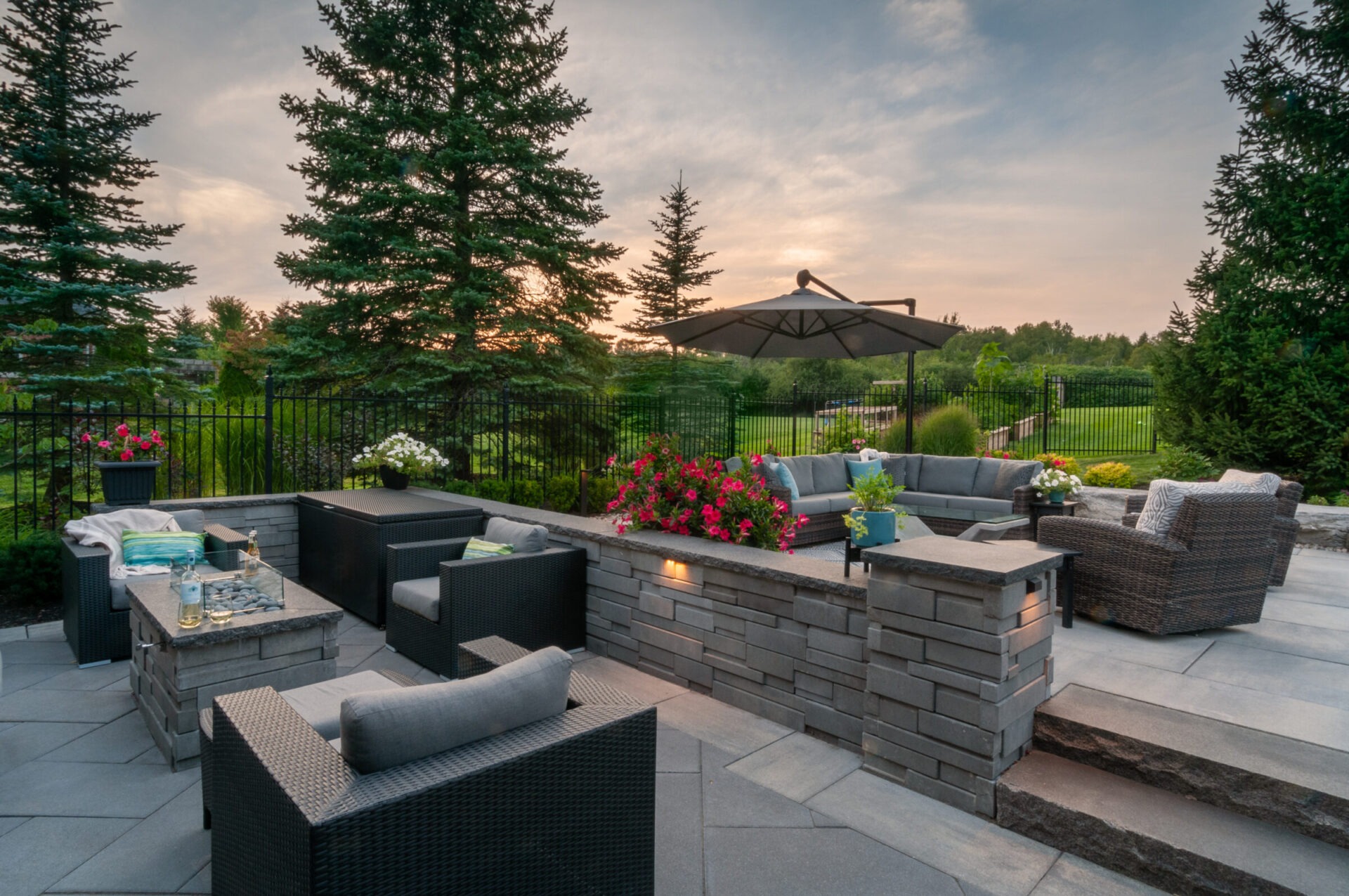 Cozy outdoor patio with wicker furniture, fire pit, and vibrant flowers, set against a backdrop of lush greenery and picturesque sunset.