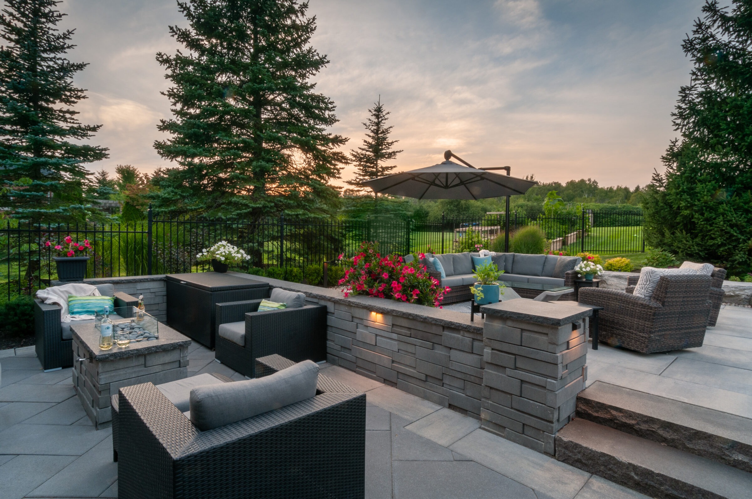 Spacious patio with modern wicker furniture, vibrant flowers, large umbrella, surrounded by trees, overlooking a serene, fenced garden at sunset.