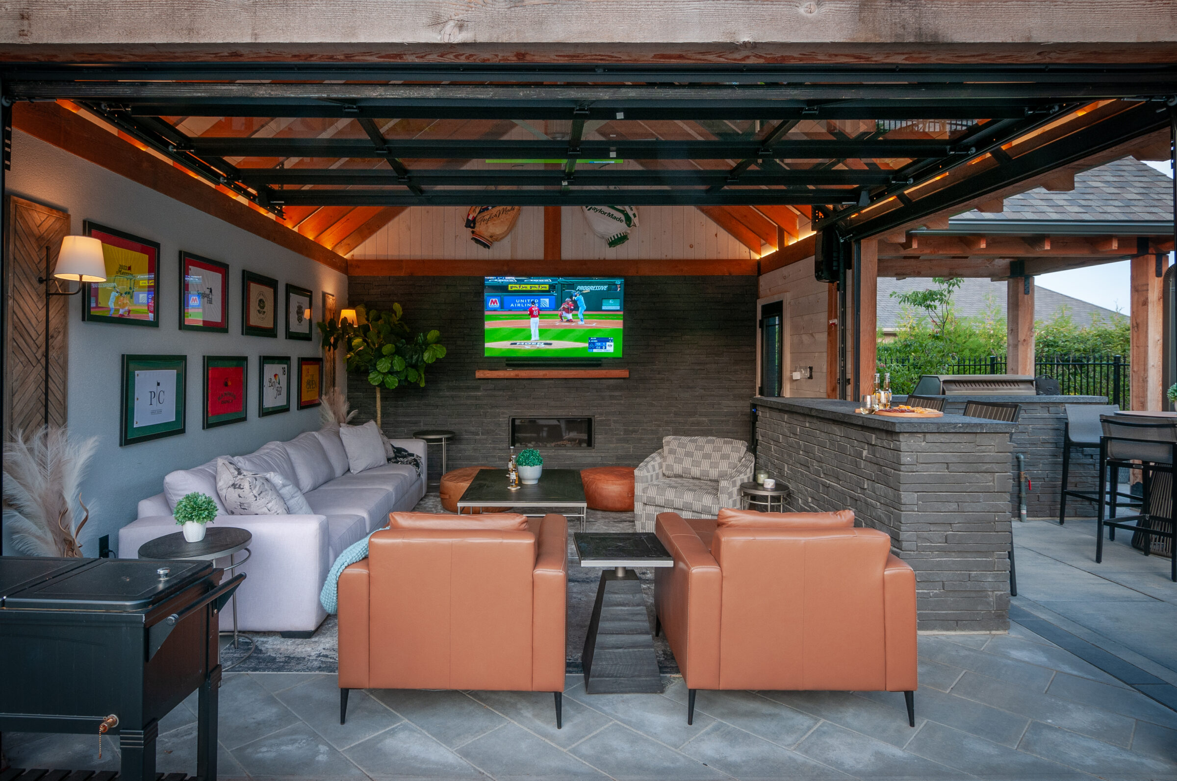 Outdoor living area with modern decor, a large TV displaying a baseball game, comfortable seating, and a bar area with stools.