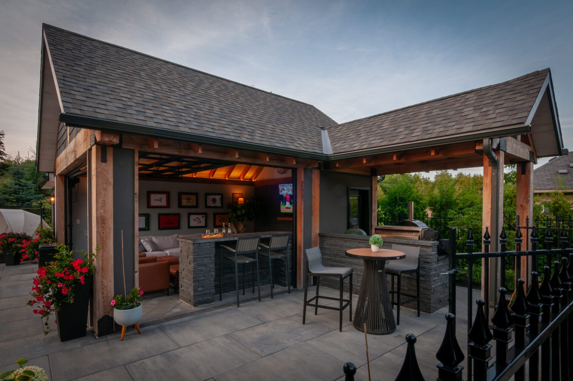 Cozy outdoor patio with a sheltered lounge area, bar seating, and vibrant flowers, under a twilight sky. Modern design with natural elements.