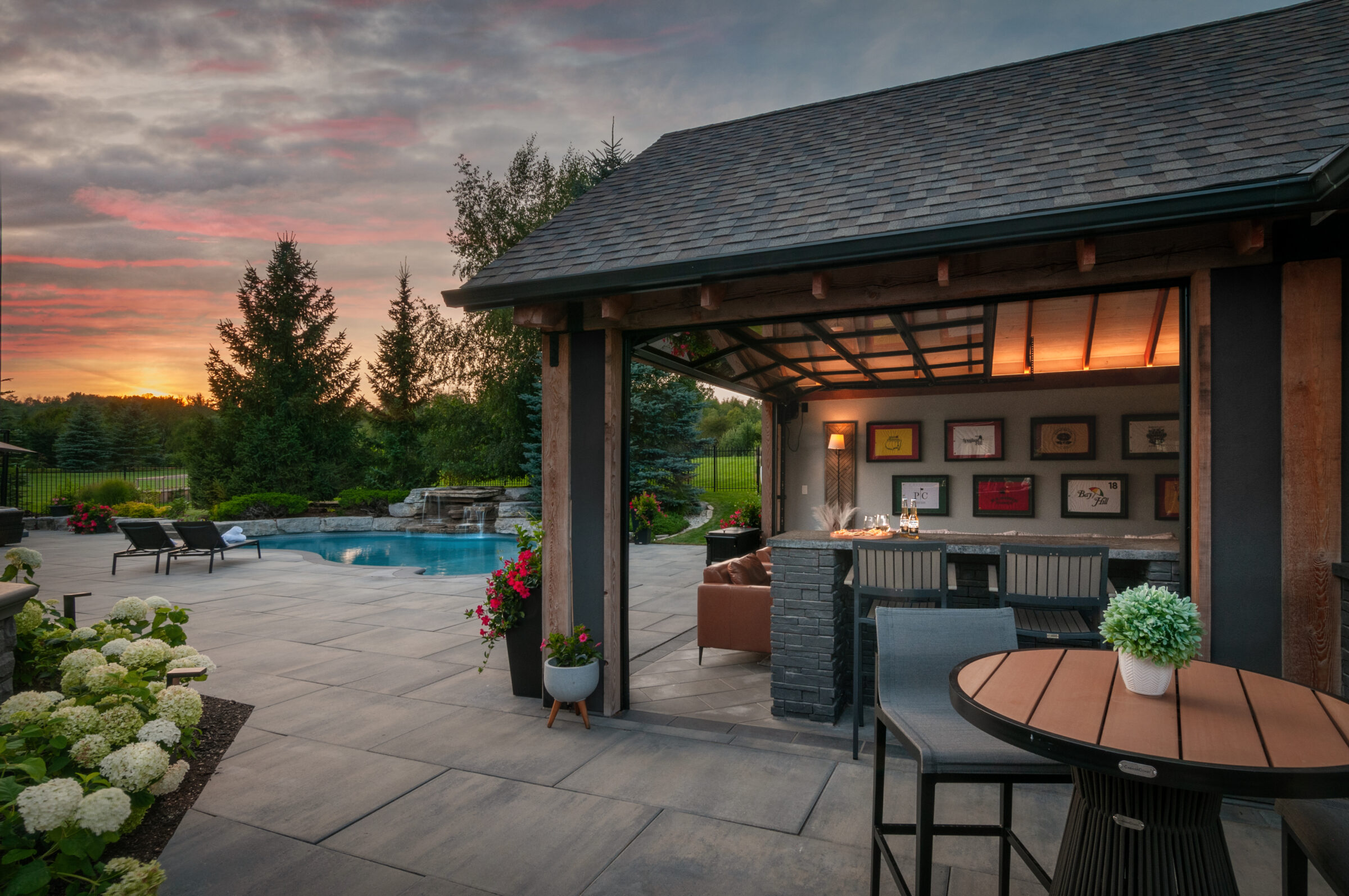 A cozy outdoor patio with a pool at sunset, featuring seating, plants, and a small bar area with framed art on the walls.