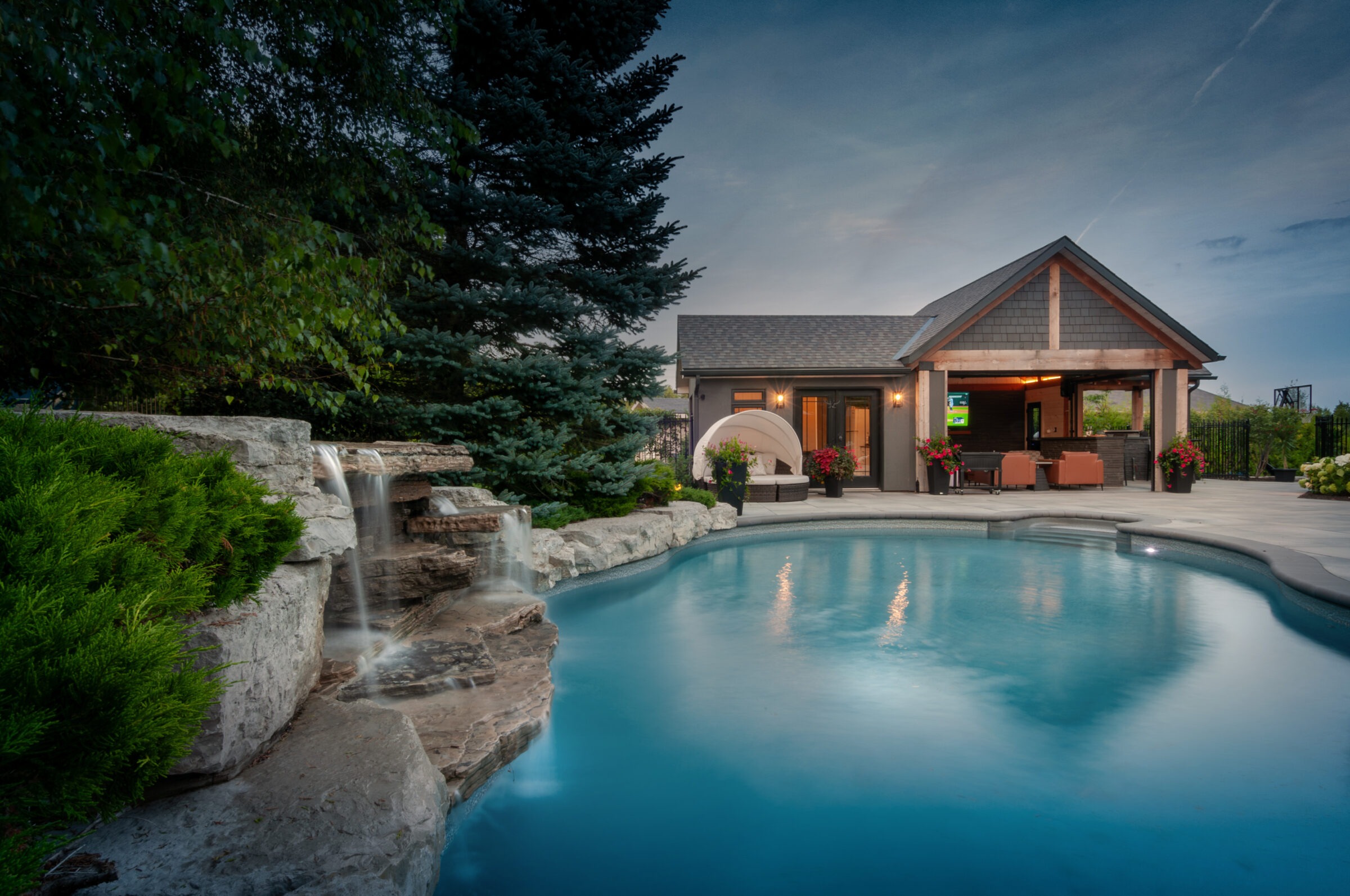 A serene backyard with a swimming pool, waterfall, lush greenery, and a cozy patio with furniture under a wooden pavilion at twilight.