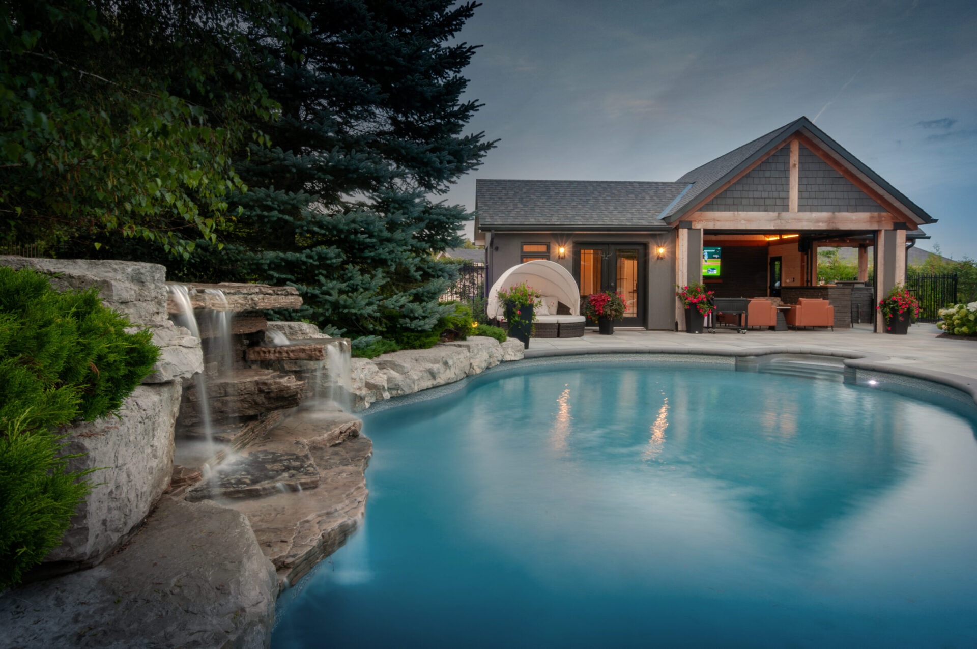 A serene backyard features a swimming pool with cascading waterfall, surrounded by lush greenery, cozy seating, and a modern pool house.