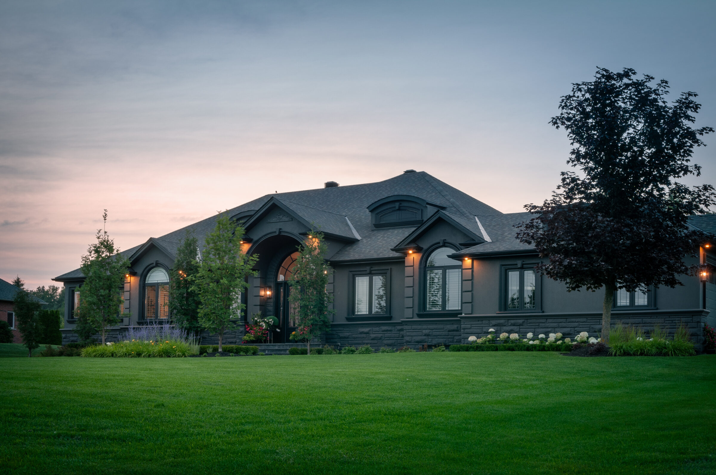 A modern, single-story house with well-maintained garden, illuminated by outdoor lights at dusk, surrounded by trees under a pinkish-blue sky.
