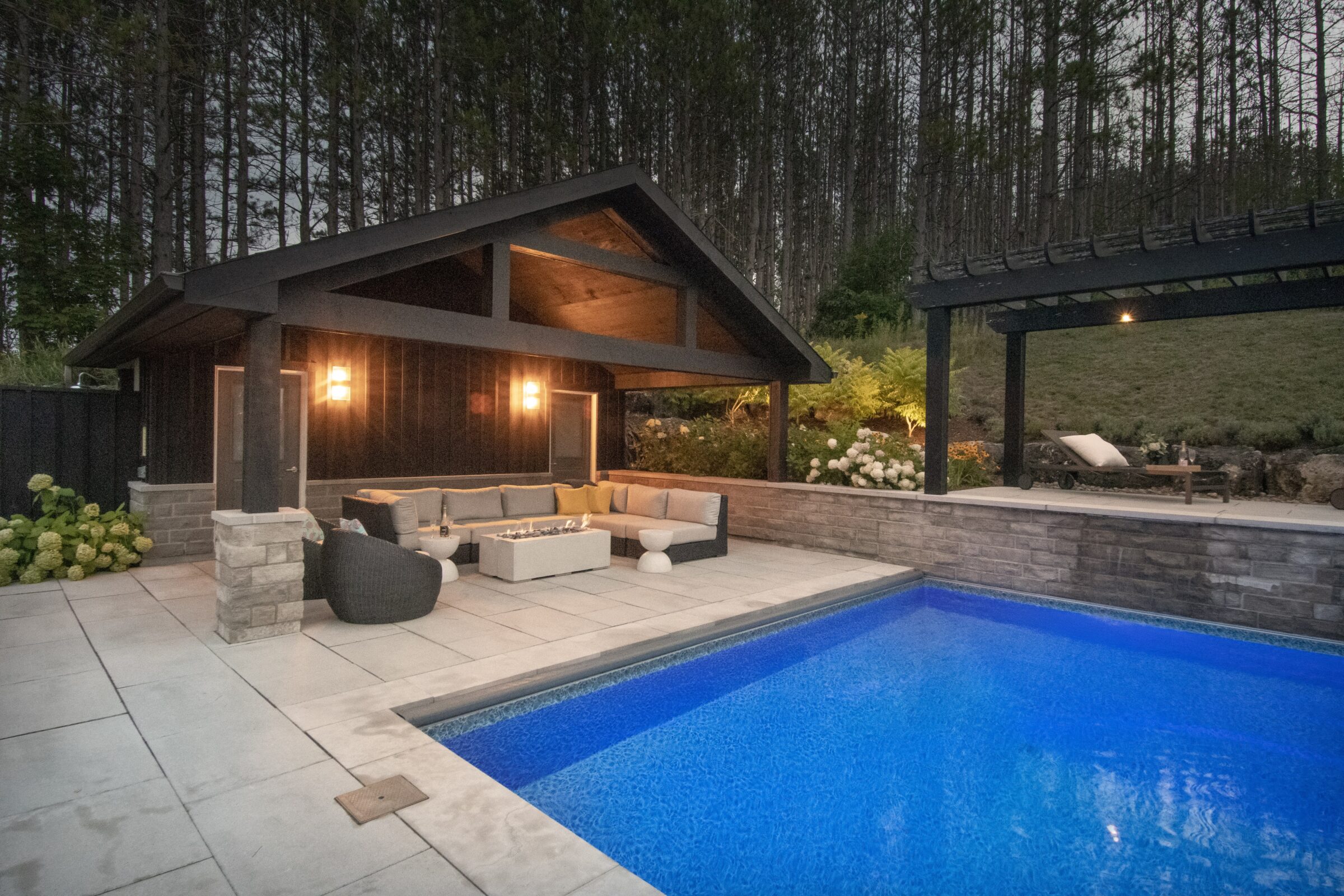 Modern backyard setting with a lit poolside cabana, cozy furniture, and surrounding trees. Elegant outdoor relaxation area with ambient lighting.