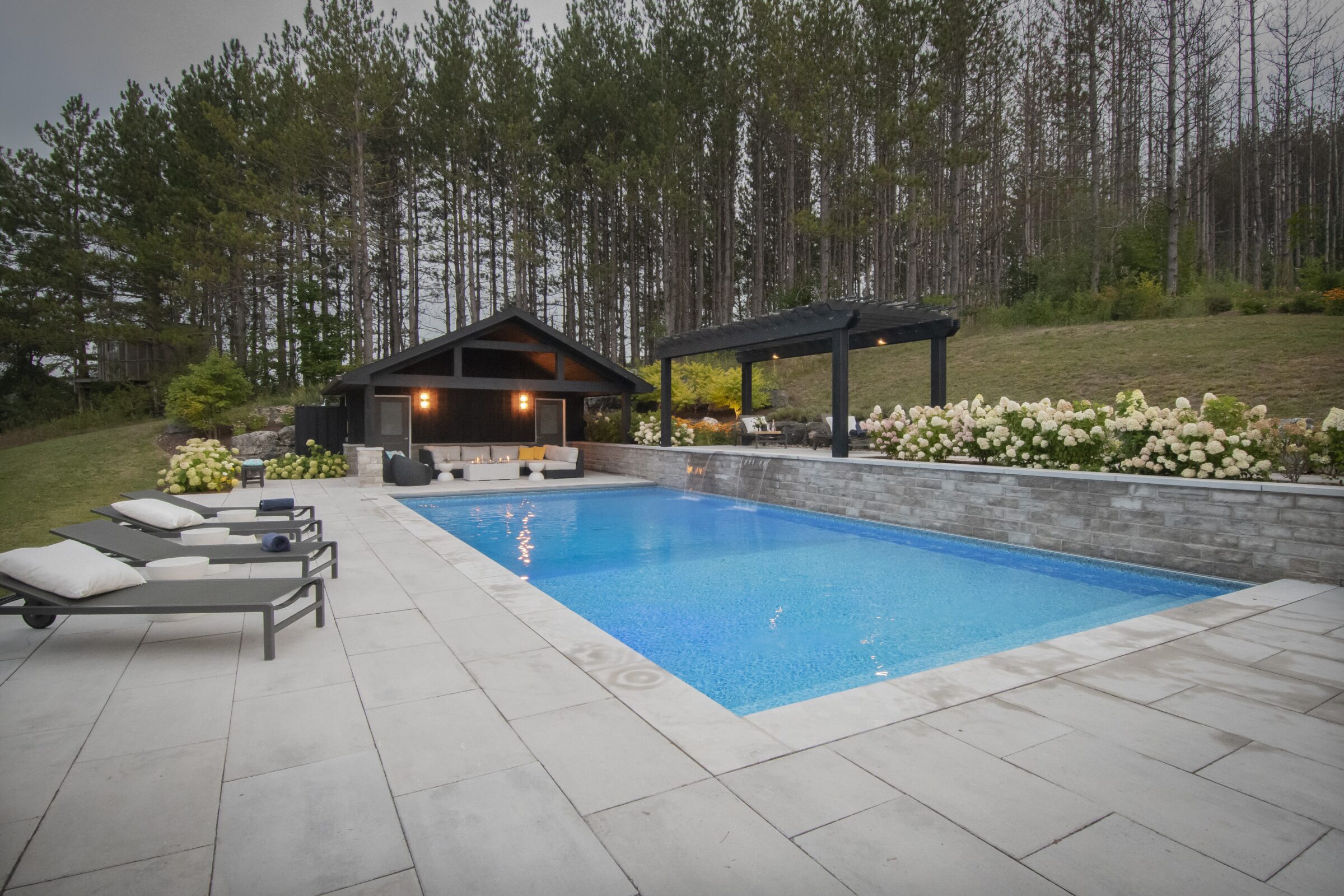 Luxurious poolside scene with loungers, modern cabana, and pergola surrounded by lush greenery and woods; peaceful, inviting outdoor retreat.