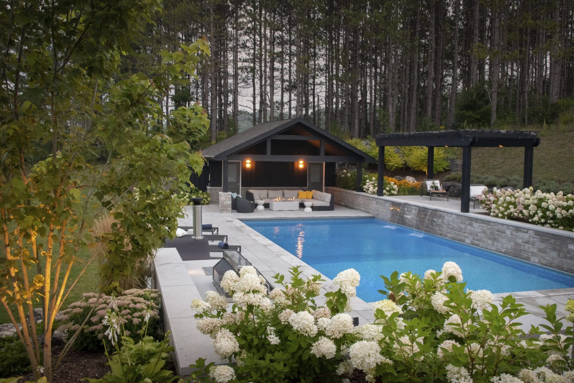 A serene backyard features a sparkling pool, cozy seating area under a pavilion, surrounded by lush greenery and elegant flowering plants.