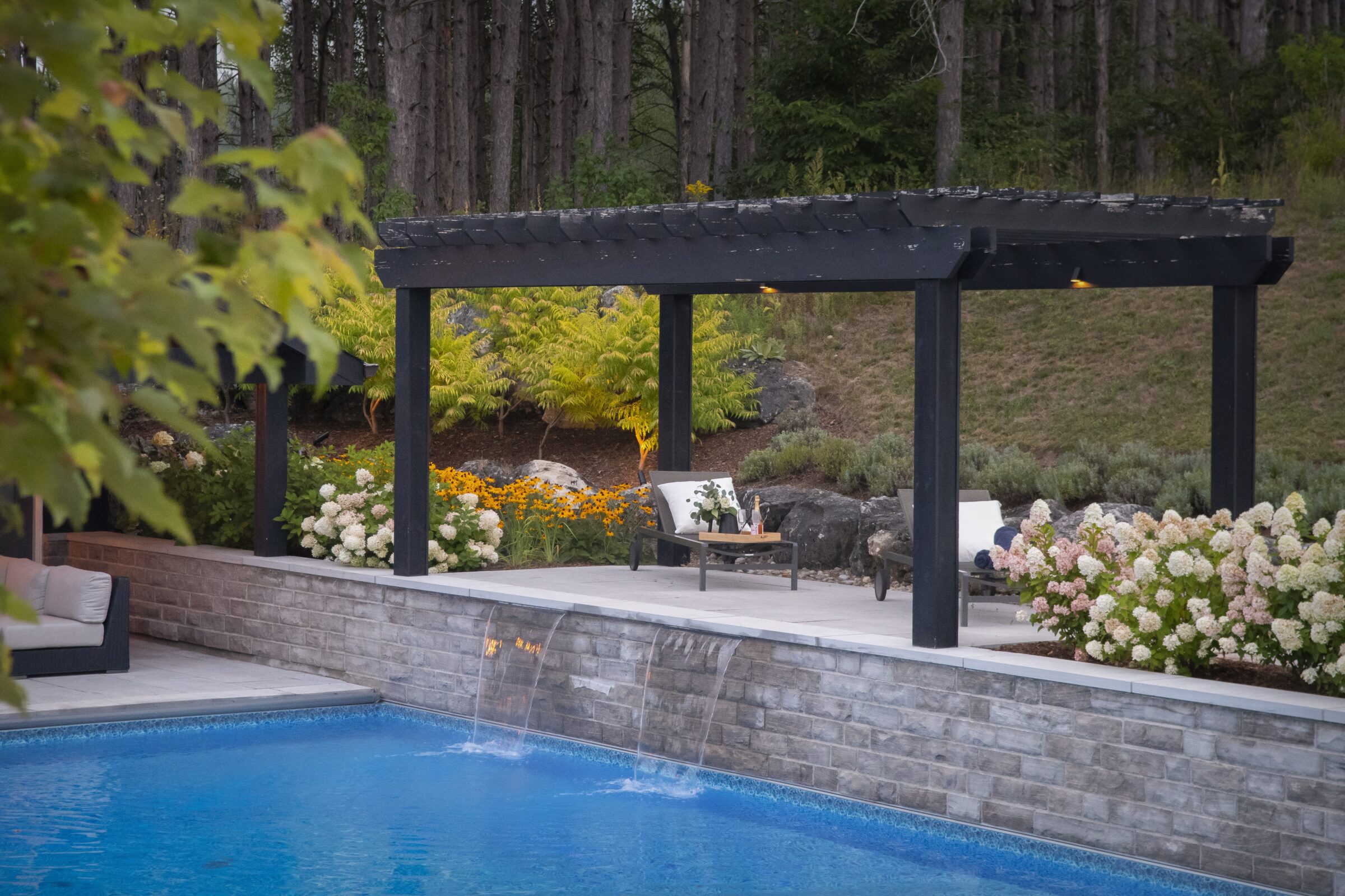A serene backyard setting with a pergola, vibrant flowers, and a flowing pool, surrounded by lush greenery and forested background.