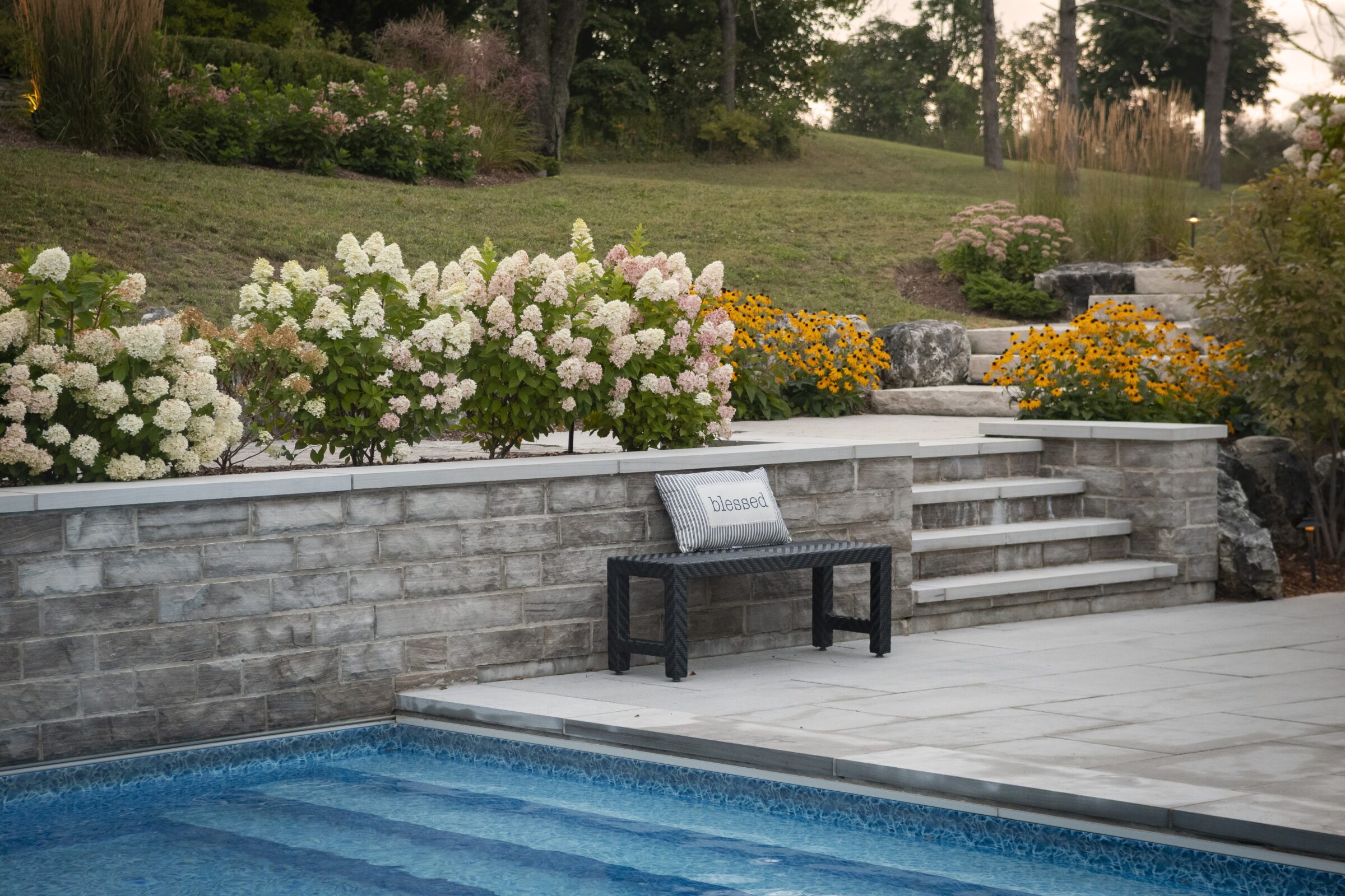 A peaceful garden with stone steps, flowers, and a pool. A bench with "blessed" cushion overlooks the serene setting.
