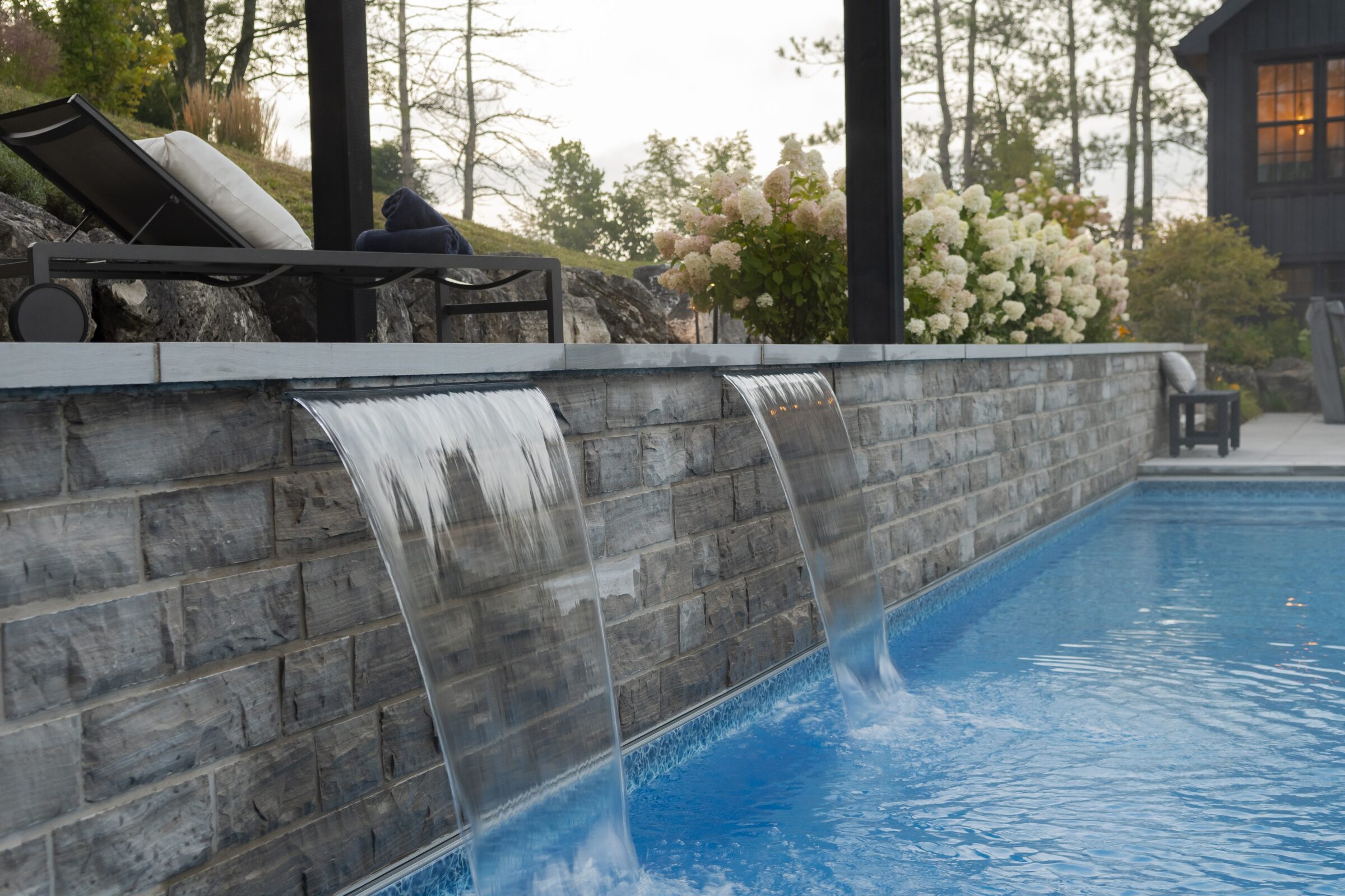 A serene outdoor pool features cascading water with lounge chairs, surrounding greenery, and flowers, creating a tranquil, relaxing atmosphere.