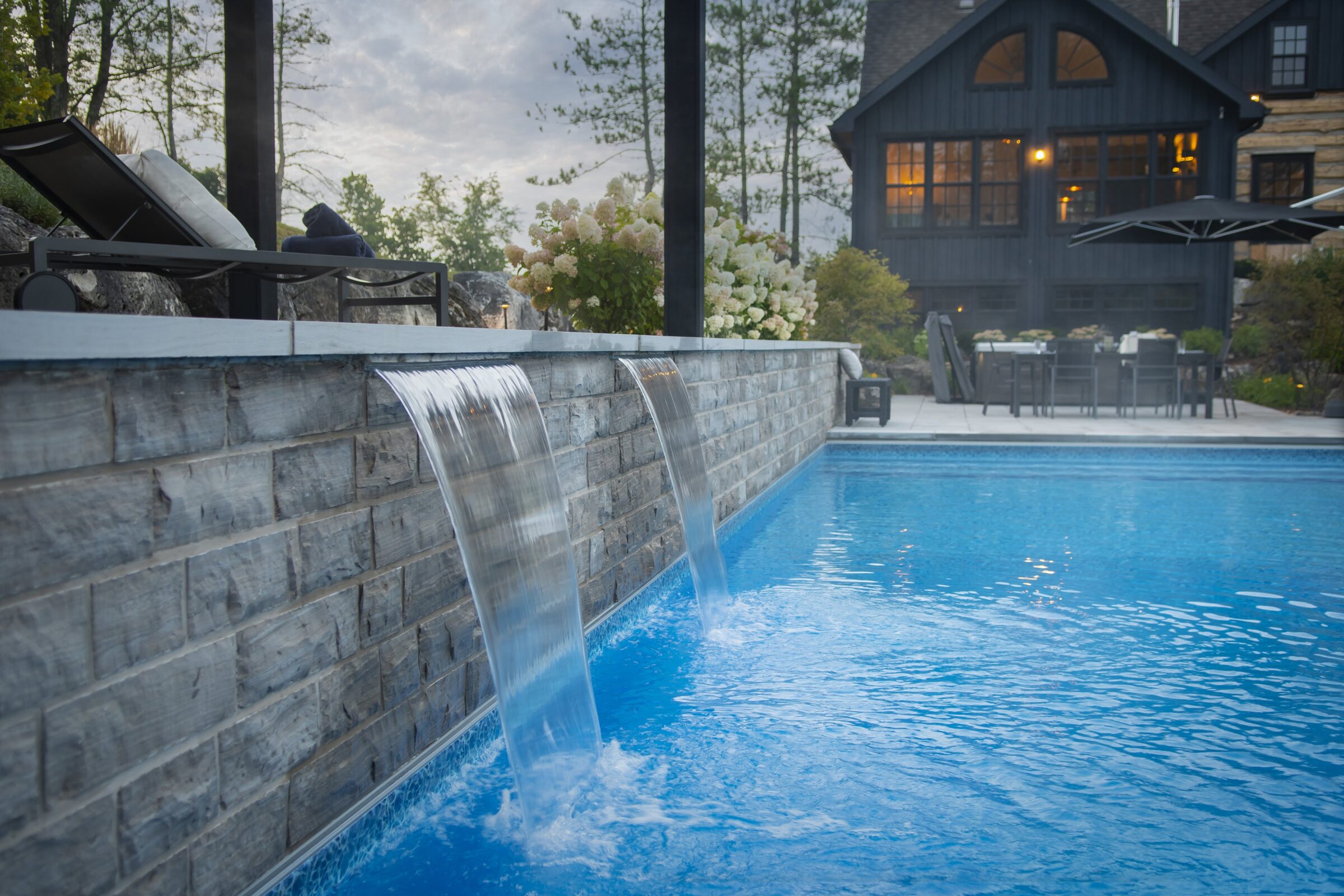 A luxurious pool with cascading waterfalls, surrounded by lounge chairs, flowers, and a modern house in the background under a cloudy sky.