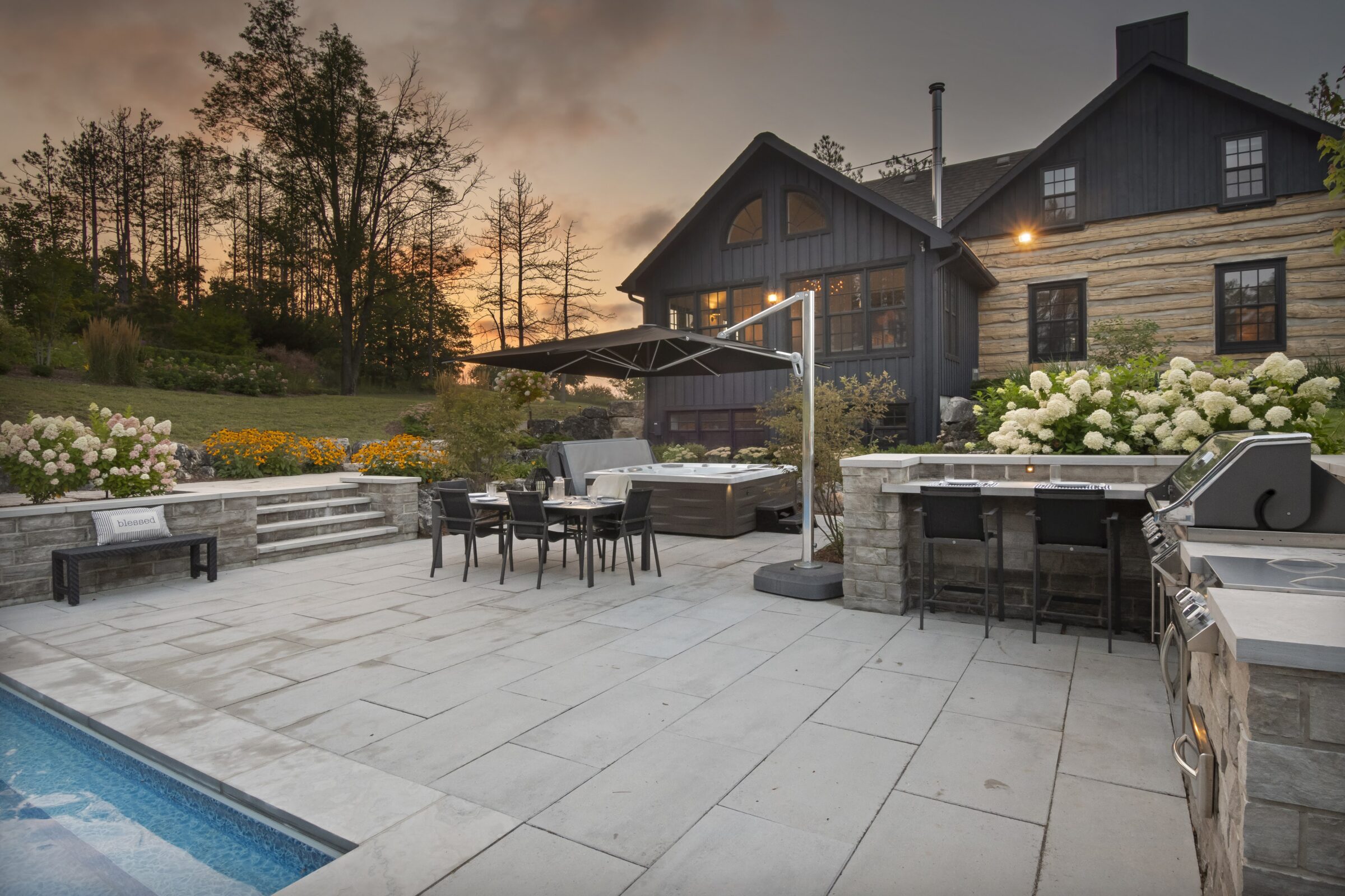 Elegant patio with a pool, outdoor dining area, and rustic house. Surrounded by flowers and trees, creating a peaceful, scenic atmosphere.