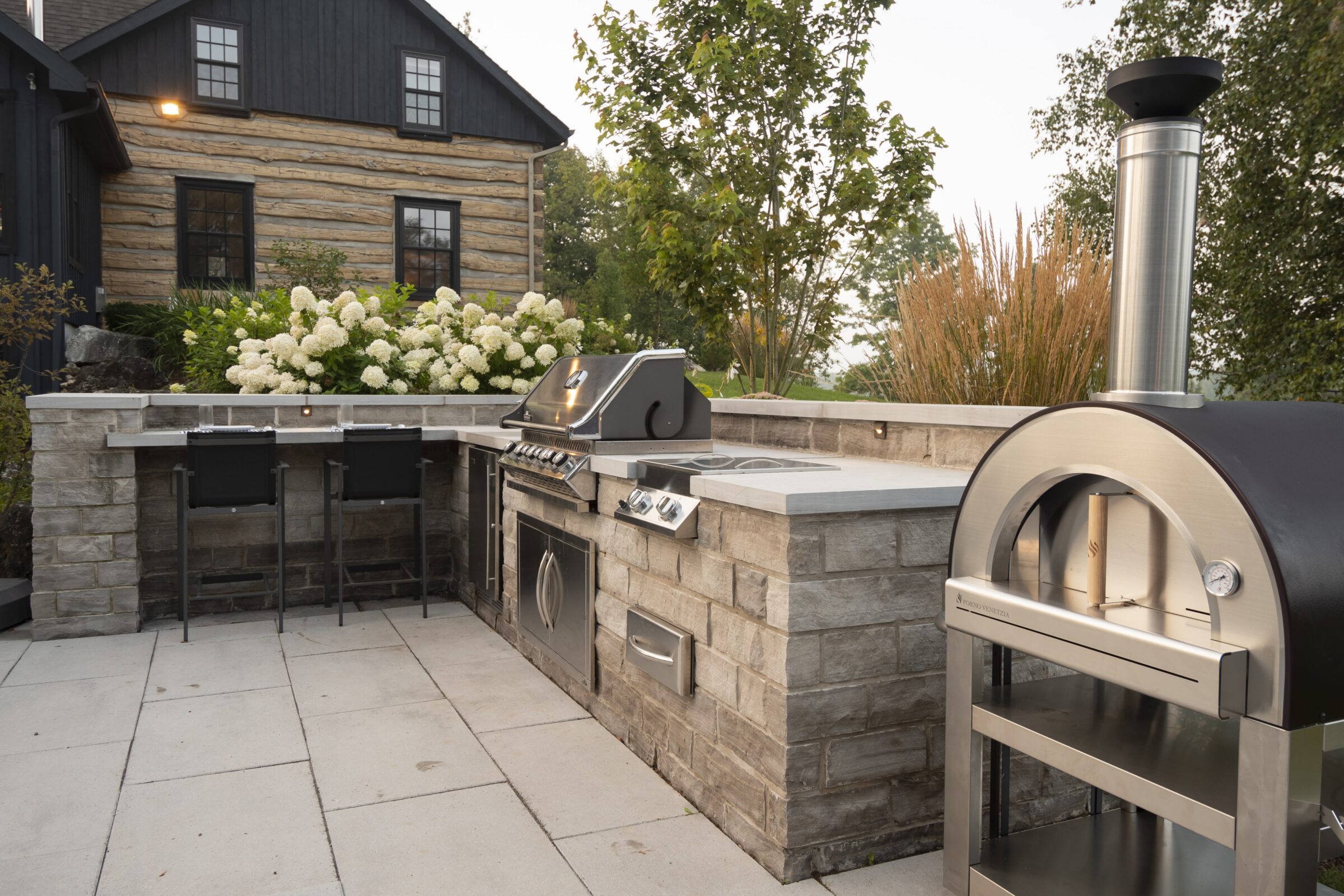 Modern outdoor kitchen with grill and pizza oven, set against a rustic house with timber details, surrounded by lush greenery and white flowers.