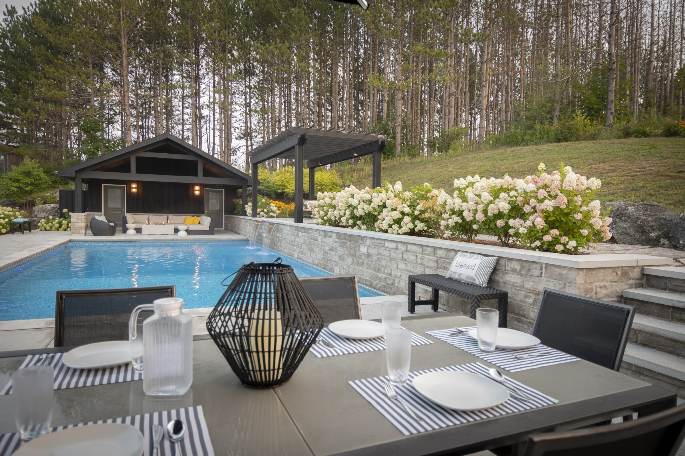A serene backyard setting features a modern patio dining area, pool, pergola, blooming hydrangeas, cozy seating, and lush pine trees in the background.