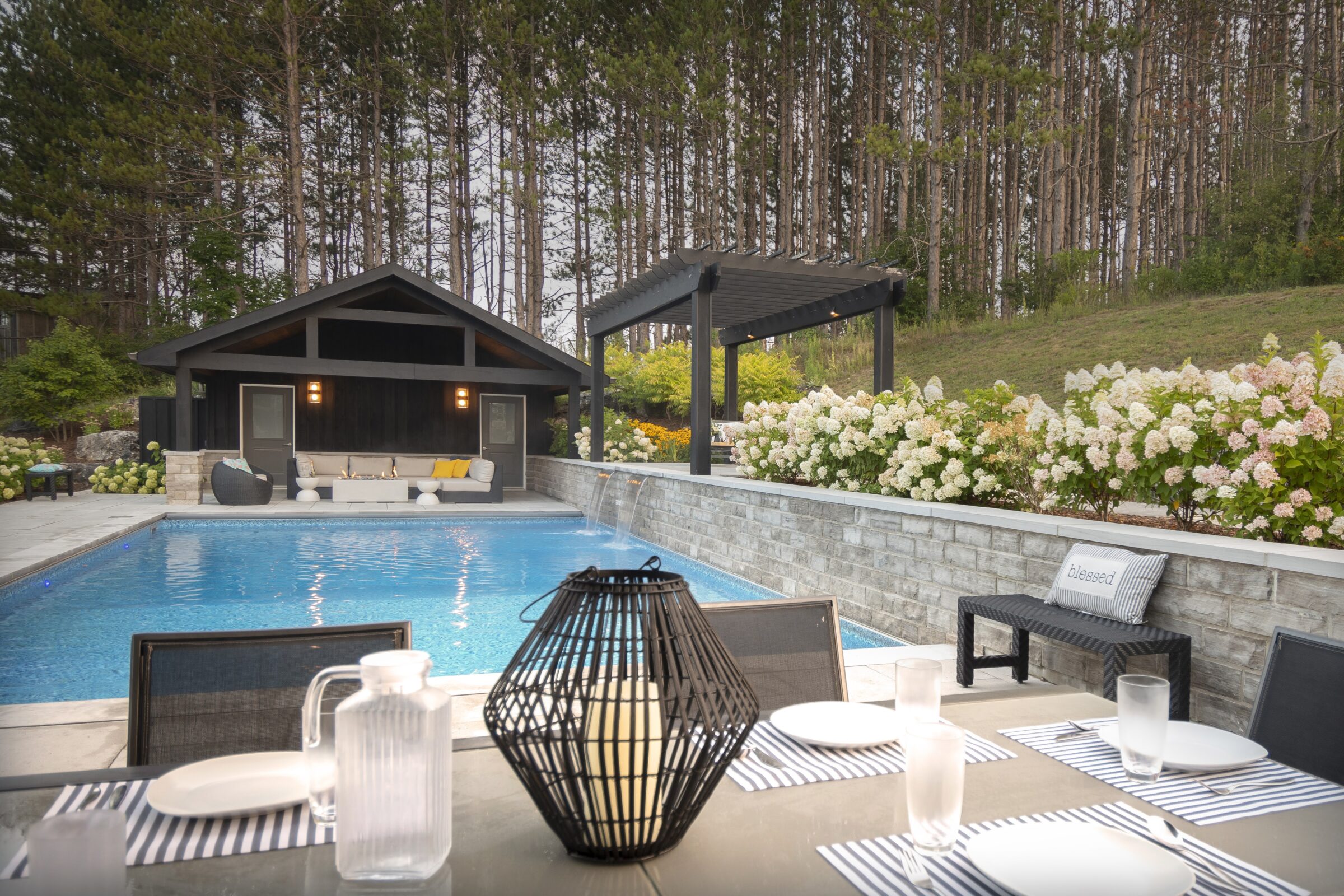 A serene backyard scene with a pool, modern pool house, dining area, pergola, flowering bushes, and tall trees in the background.