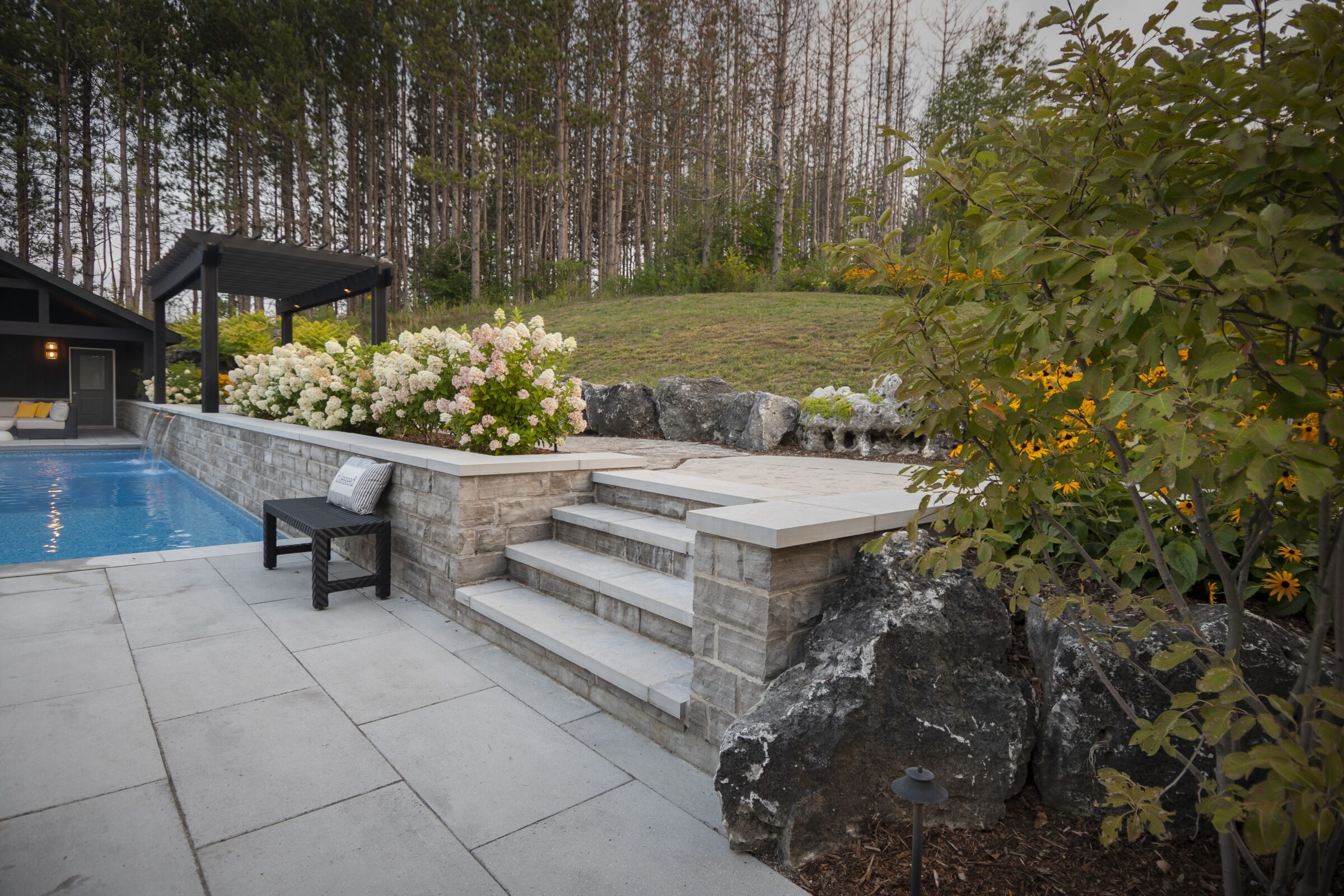 A serene backyard features a pool, patio with steps, lush flowers, and surrounding trees, creating a tranquil outdoor retreat.