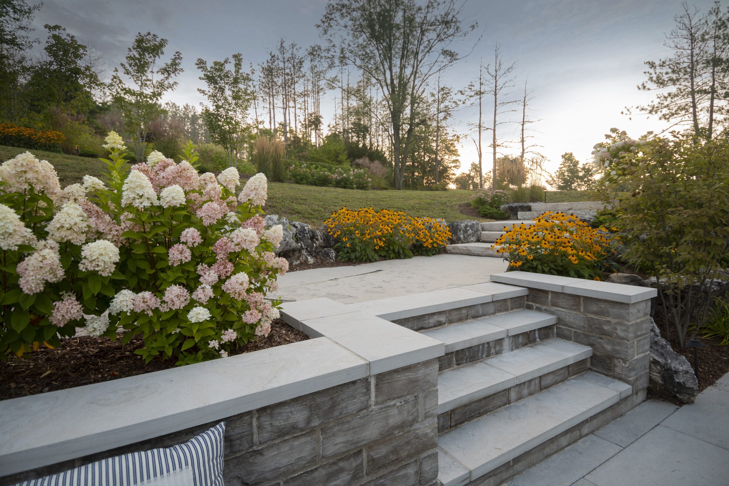 A landscaped garden with stone steps, colorful flowers, and surrounding trees under a partly cloudy sky at dusk.