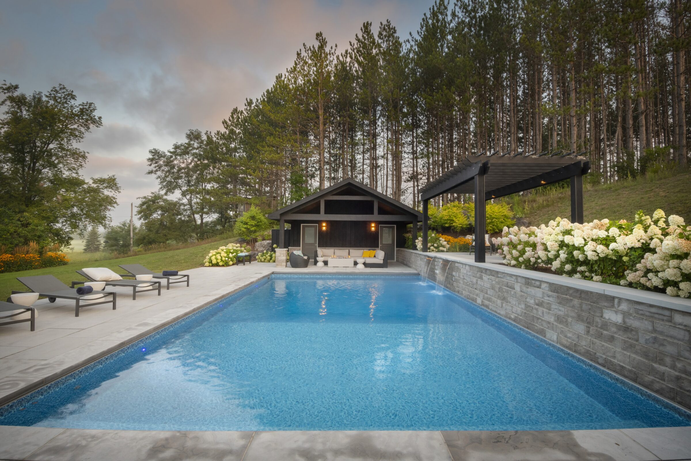 A serene outdoor pool area with lounge chairs, surrounded by lush trees and flowers, adjacent to a modern pavilion under a colorful sky.