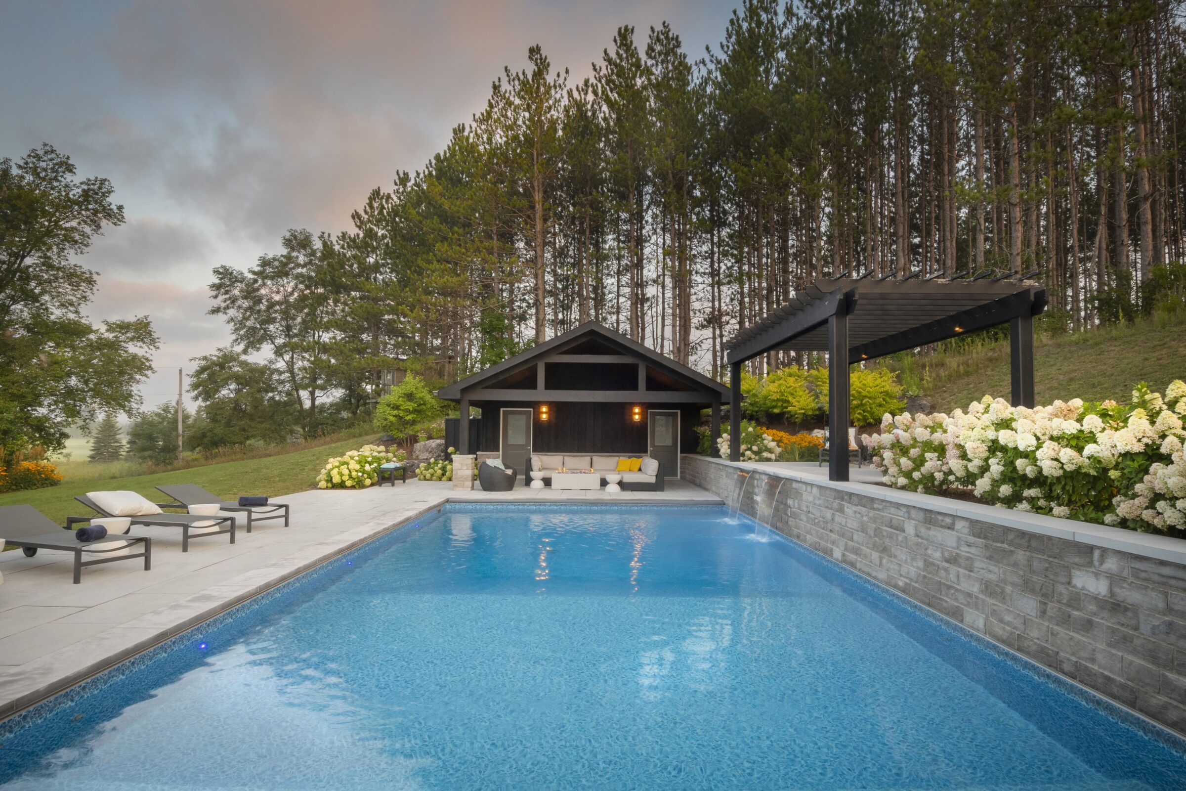 A serene poolside setting with lounge chairs, pergola, and cozy seating area, surrounded by lush greenery and tall trees under a colorful sky.