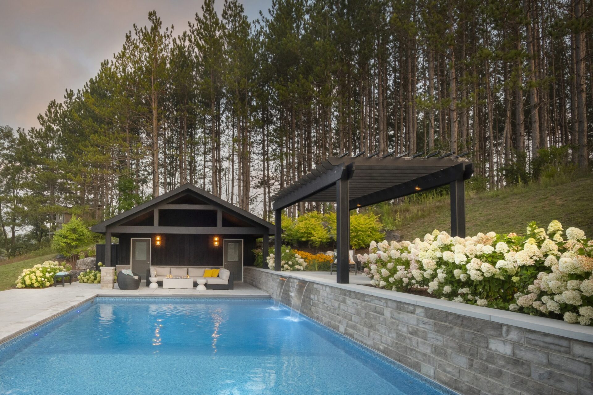A serene backyard pool with a modern patio, surrounded by lush greenery and tall trees, features a pergola and cozy seating area.