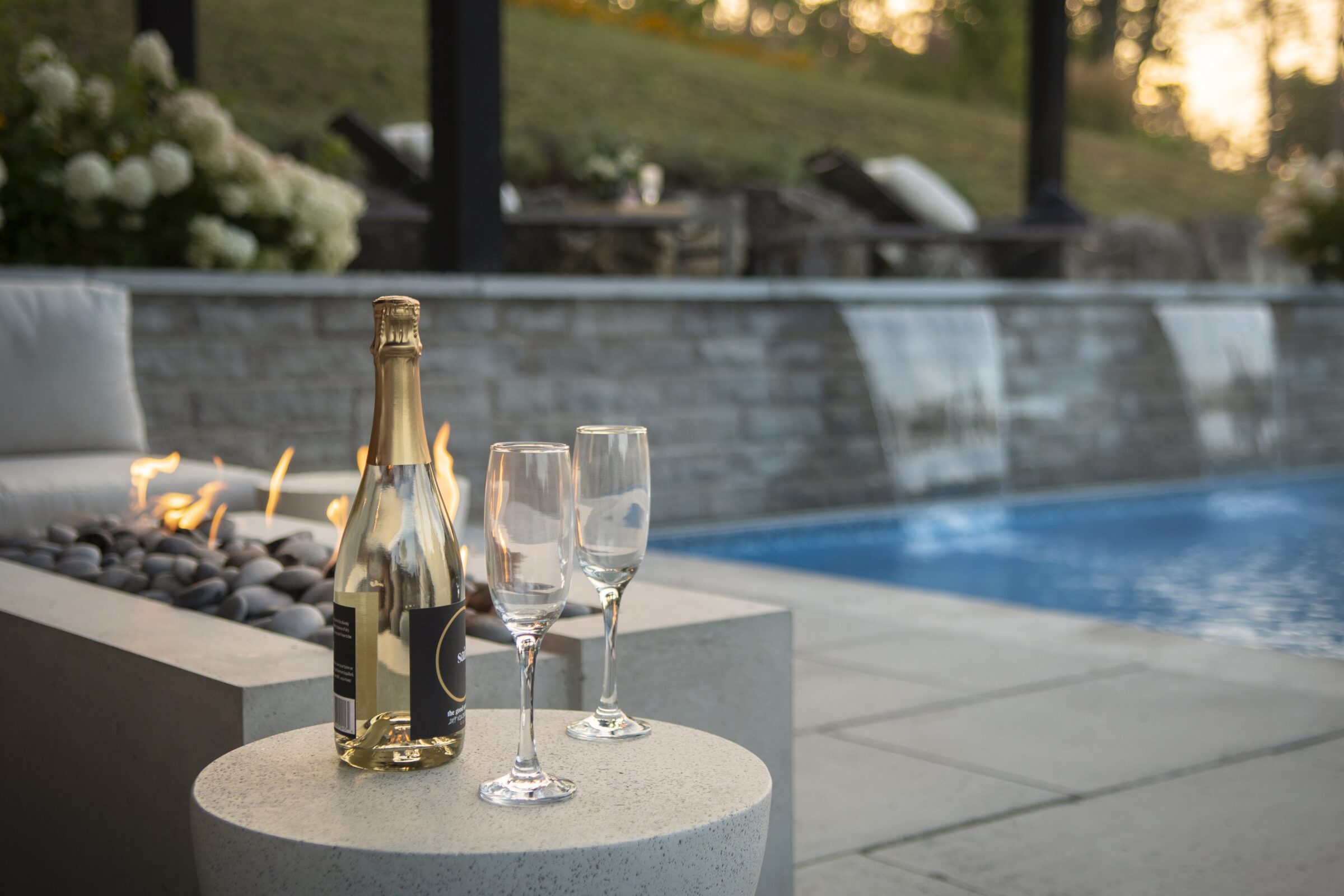 A tranquil poolside setting with a champagne bottle, glasses, fire pit, and relaxing water feature under the evening sky.