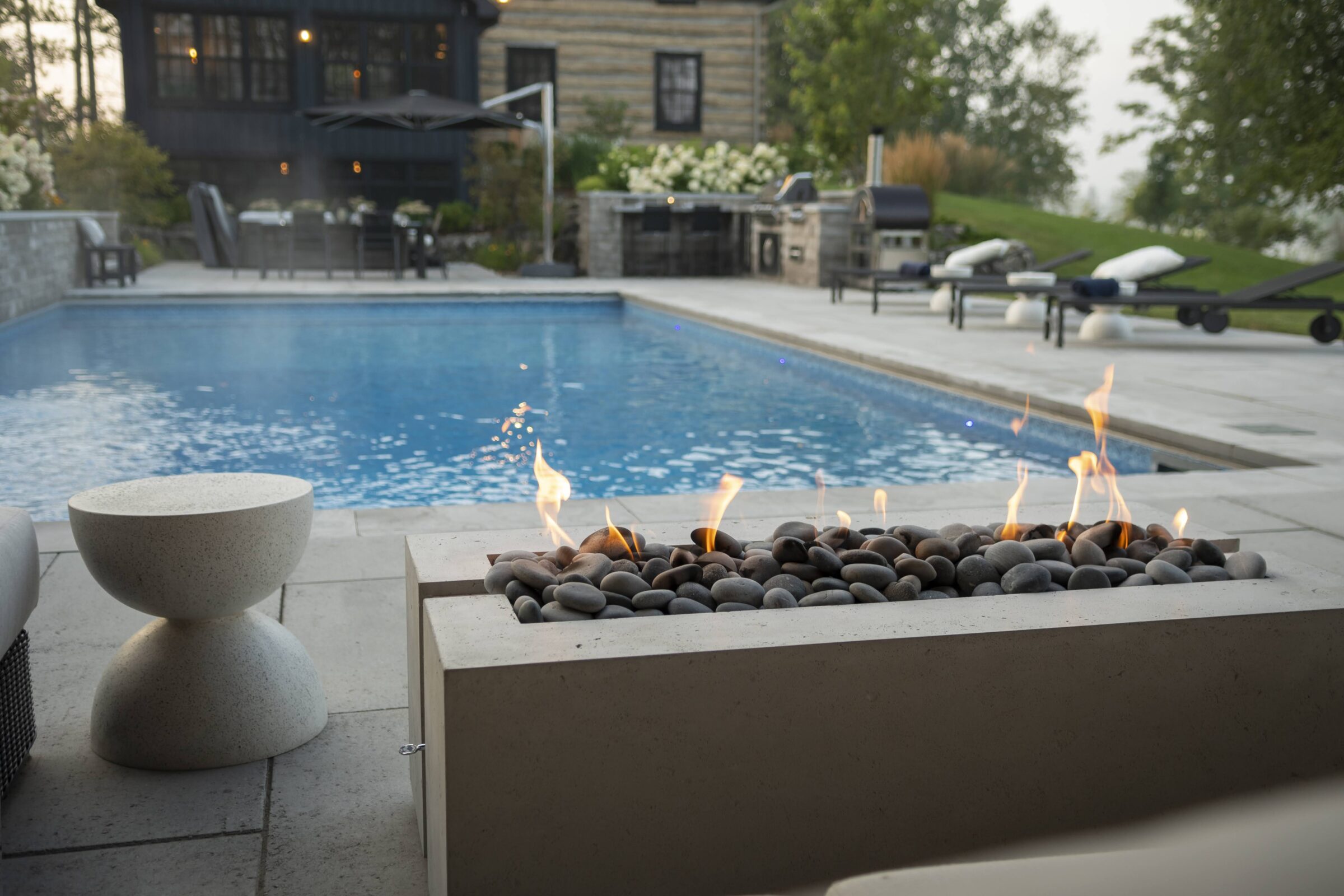 A serene poolside setting featuring a modern fire pit, lounge chairs, dining area, and lush greenery, offering a relaxing outdoor ambiance.