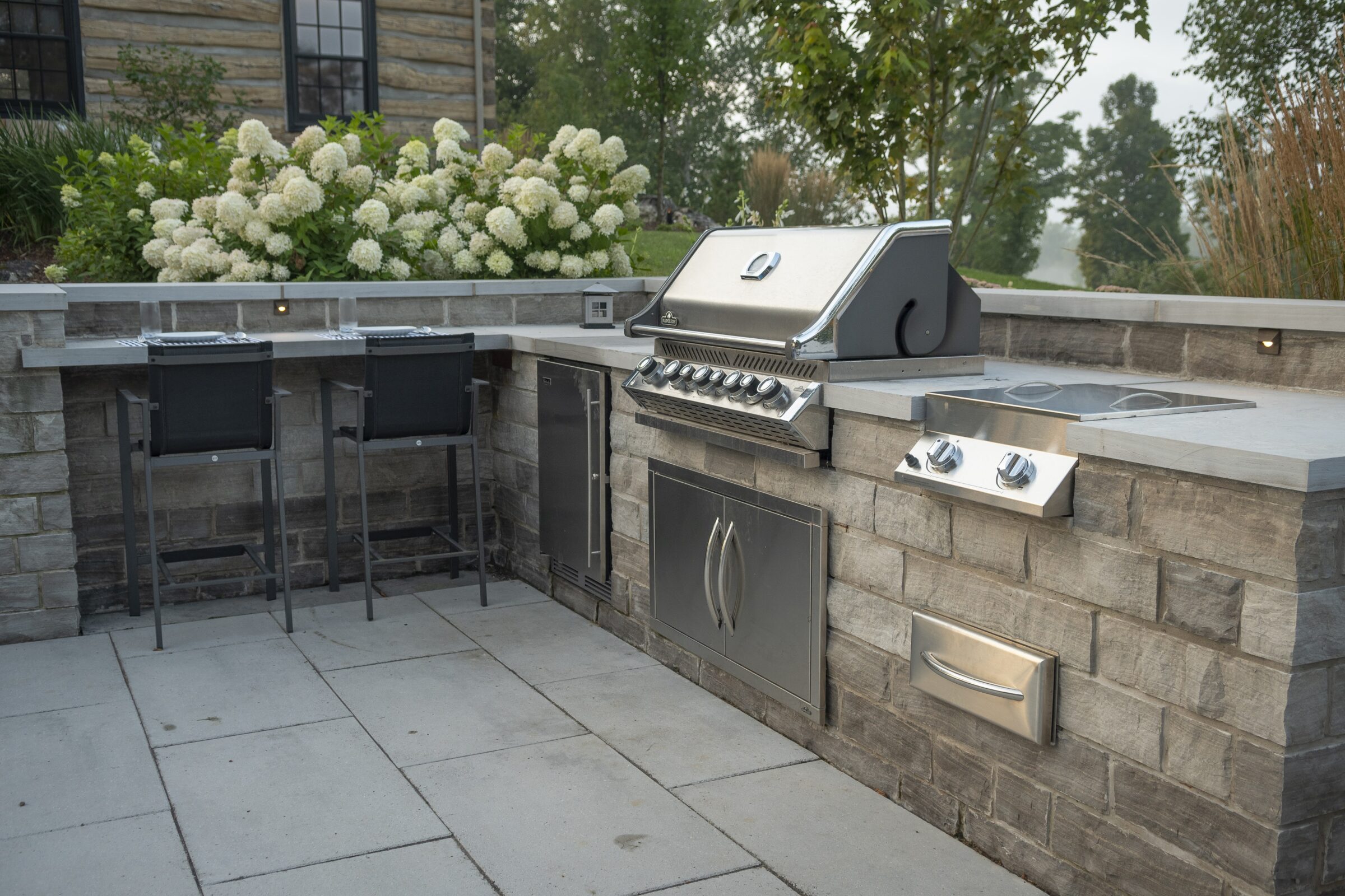 Outdoor kitchen with stainless steel grill, two barstools, and stone counter. Lush garden and wooden house exterior in the background.