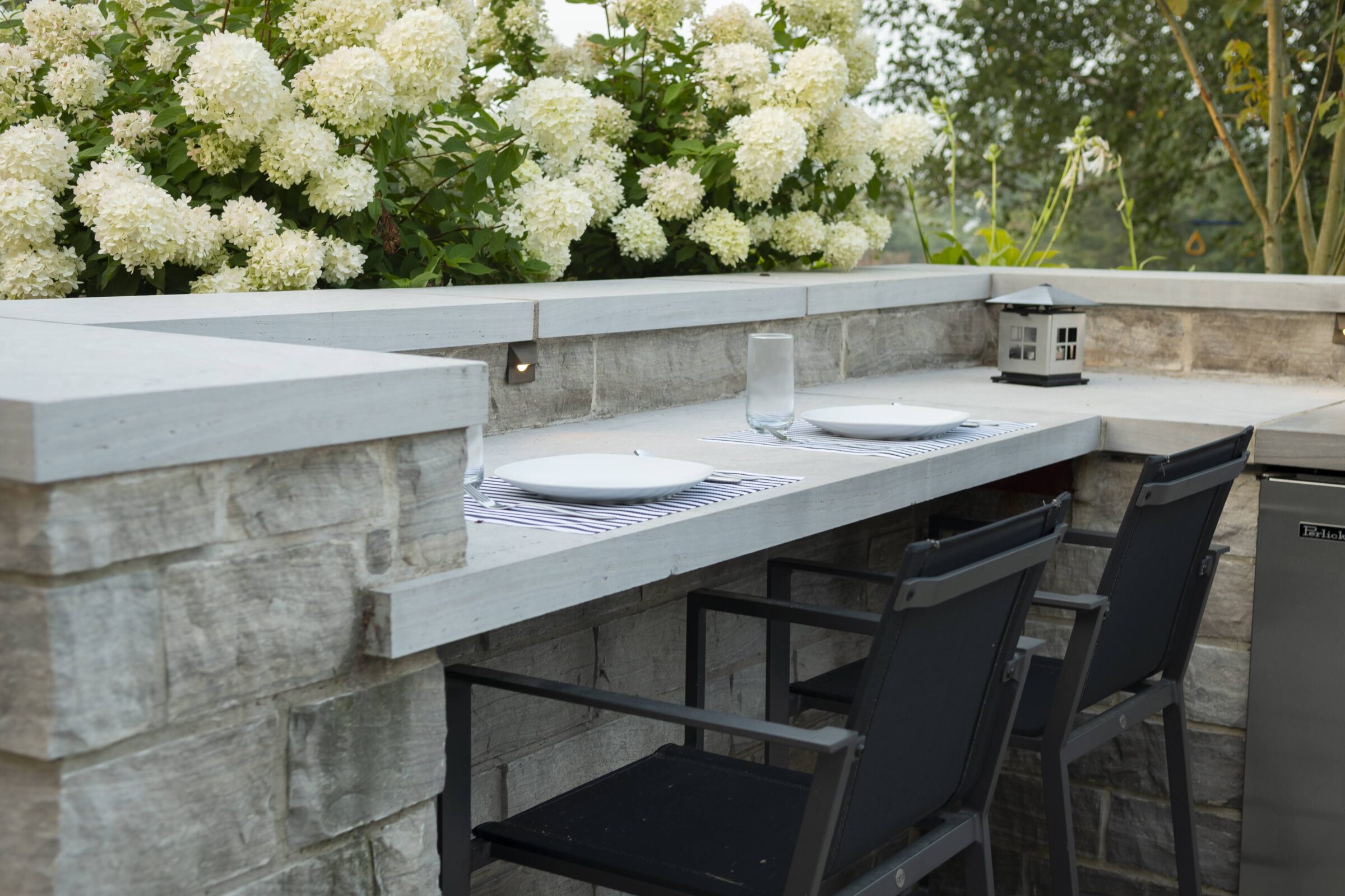 Outdoor patio setting with two chairs, a stone bar, white flowers, lantern, and table settings, creating an elegant, serene atmosphere.