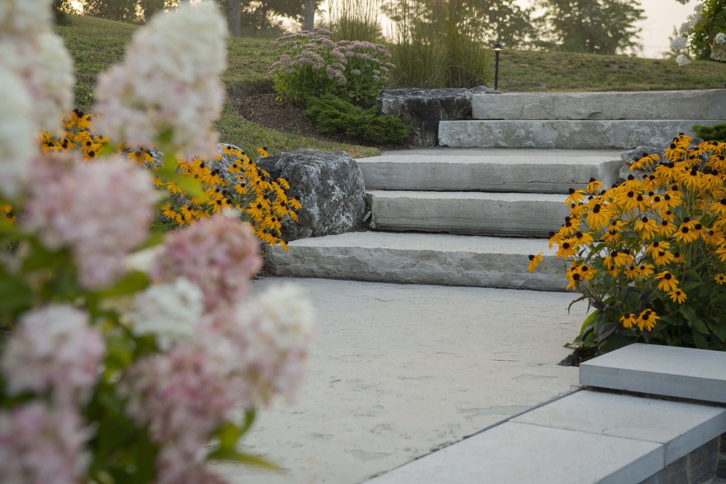 Stone steps lead through a garden with vibrant yellow and pink flowers, set in a tranquil, green landscape.