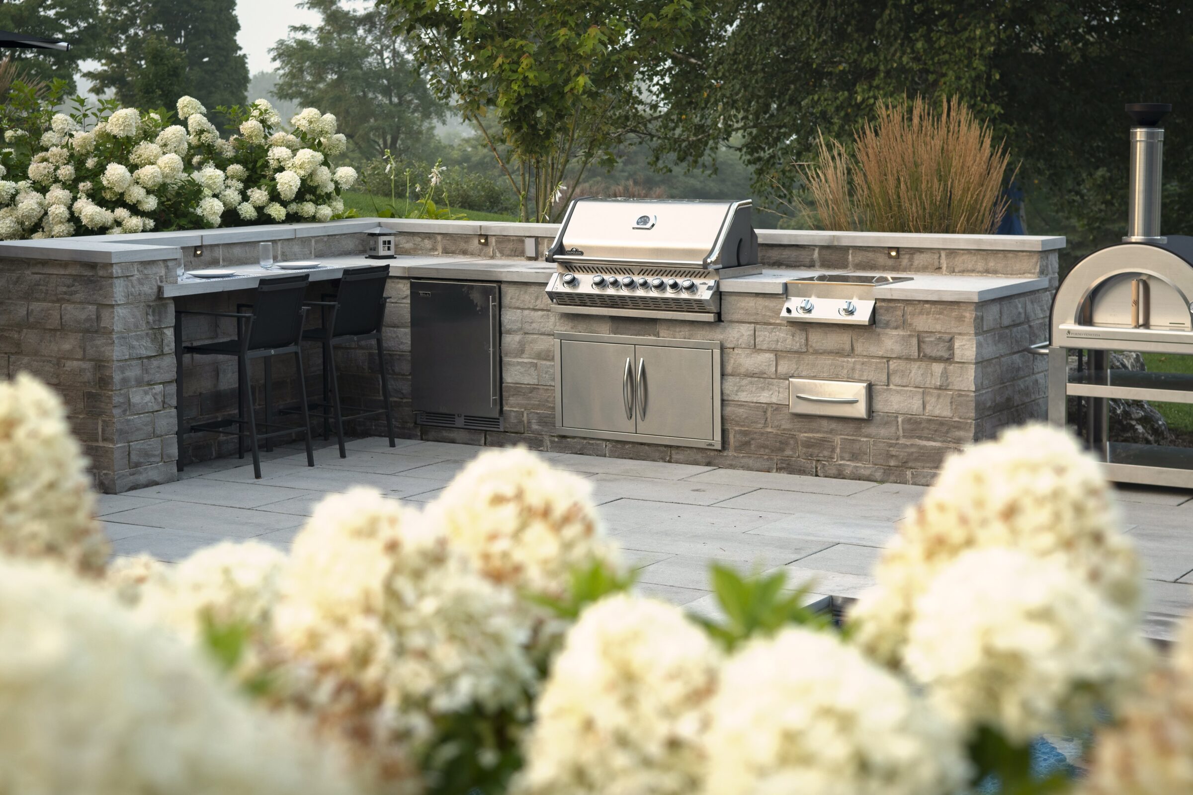 Outdoor kitchen with stainless steel grill, stone countertops, and seating. Surrounded by white flowers and greenery in a serene garden setting.
