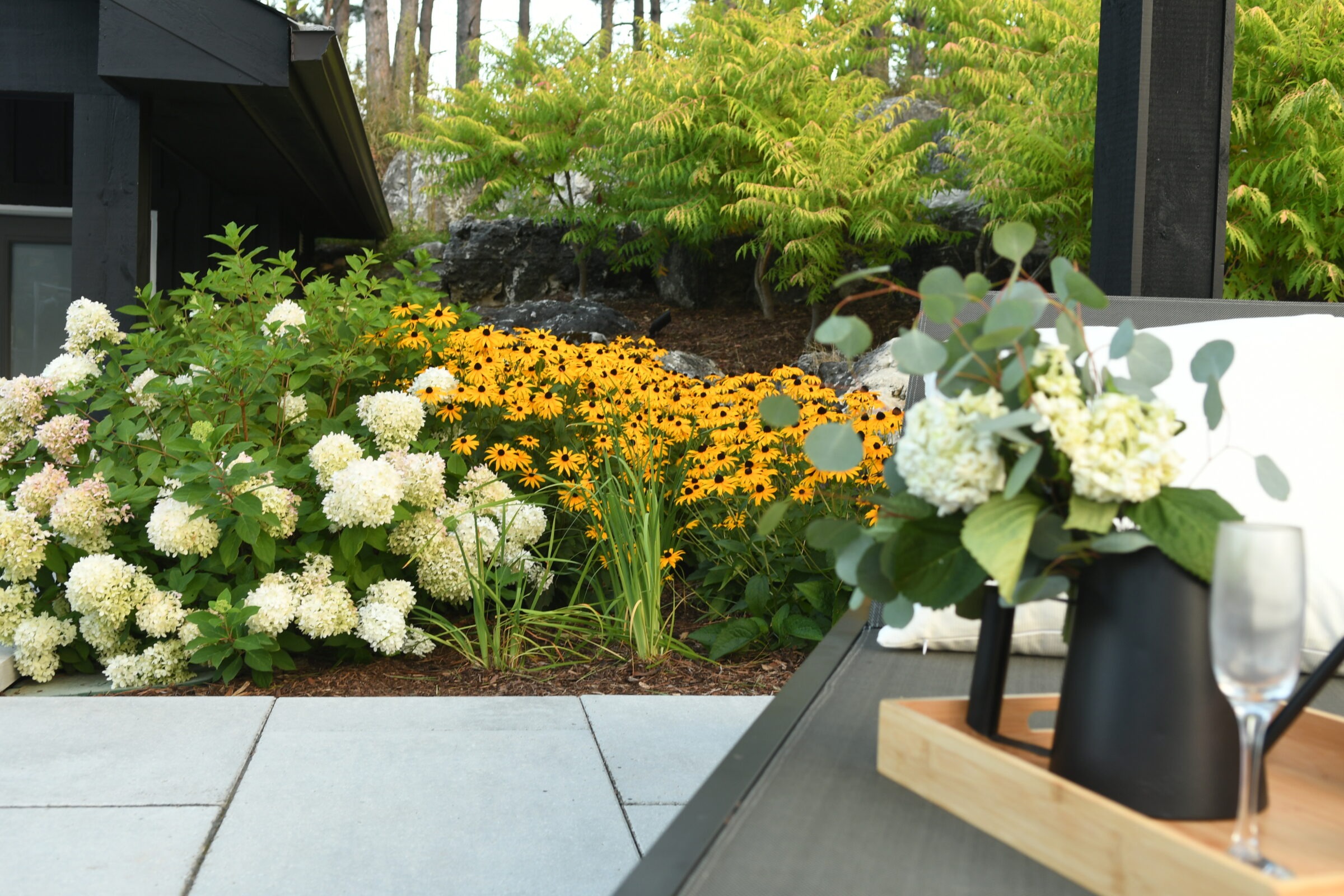 A lush garden scene features vibrant yellow daisies, white hydrangeas, and a wooden tray with flowers and a glass on a patio chair.