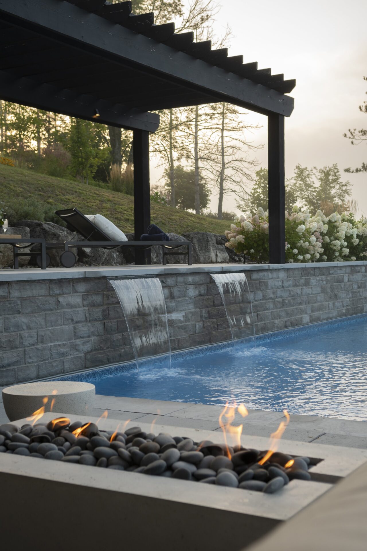 Modern poolside with a pergola, fire pit, and cascading waterfalls. Surrounding greenery provides a serene backdrop for relaxation. No people visible.