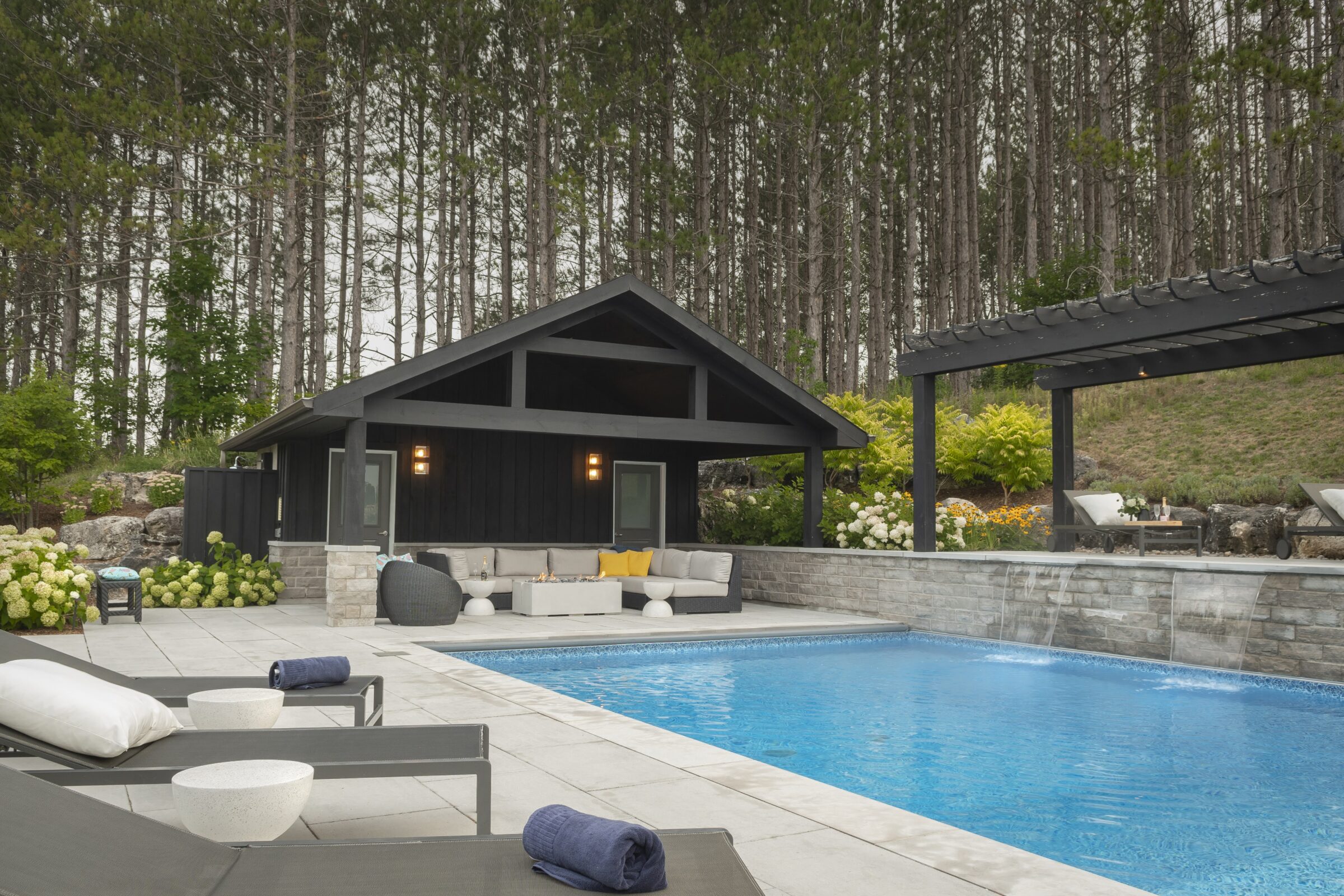 Modern poolside with loungers, a cozy seating area under a pavilion, surrounded by lush greenery and tall trees in a serene setting.