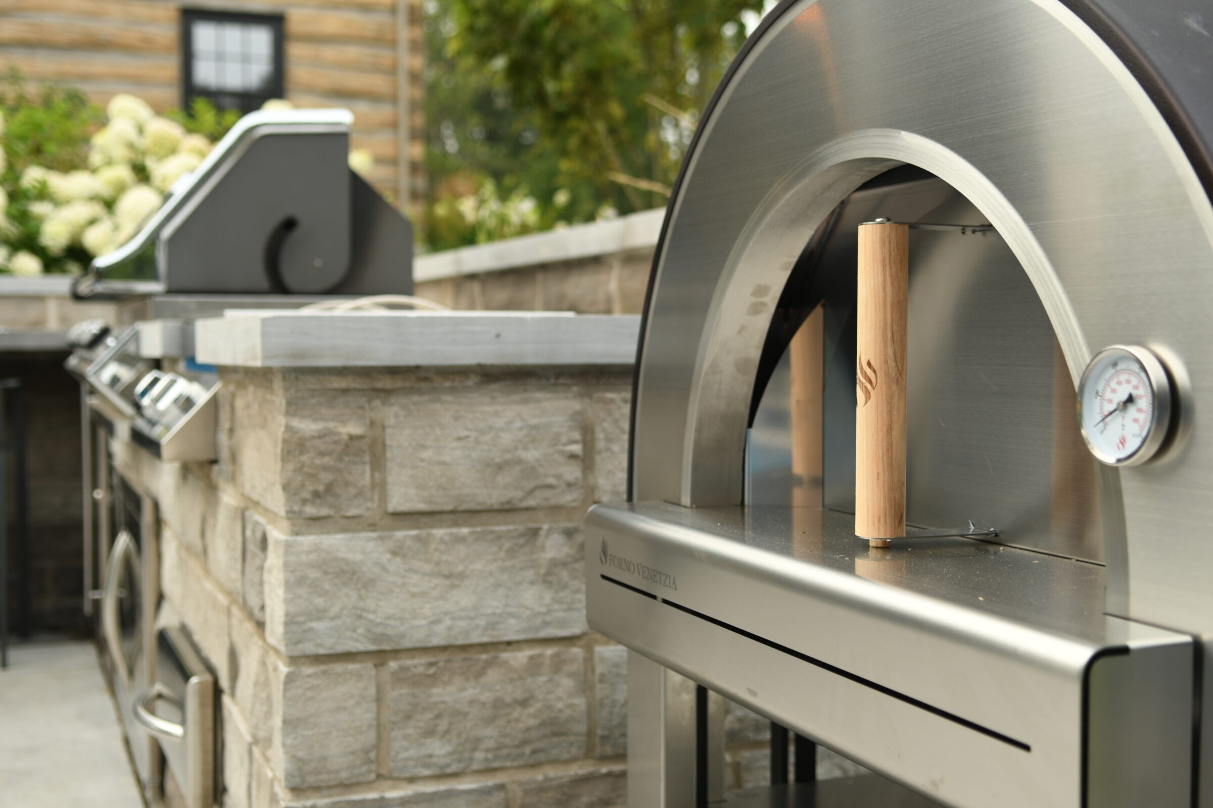 Modern outdoor kitchen featuring a stainless steel pizza oven and grill set within a stone counter, surrounded by lush greenery and flowers.