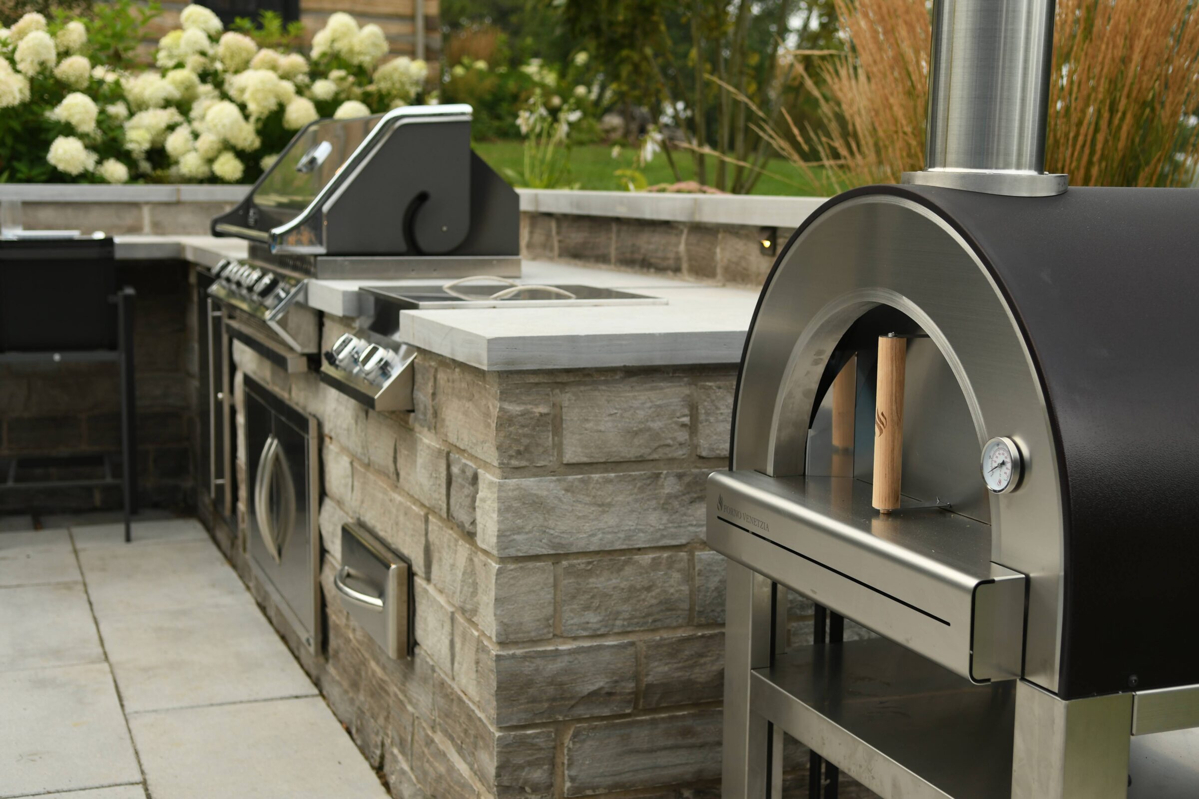 Outdoor kitchen with stainless steel barbecue grill, pizza oven, and stone countertops surrounded by green garden and white flowers. No people present.