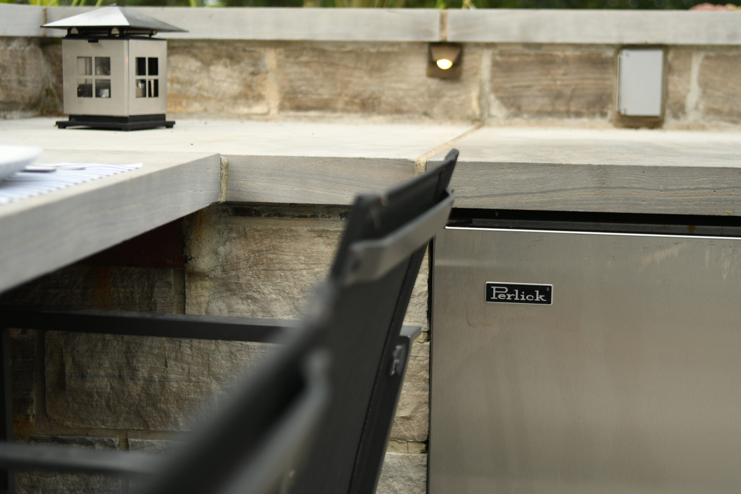 Outdoor patio scene with stone wall, metal lantern, and stainless steel Perlick appliance. Black chair partially visible in the foreground.