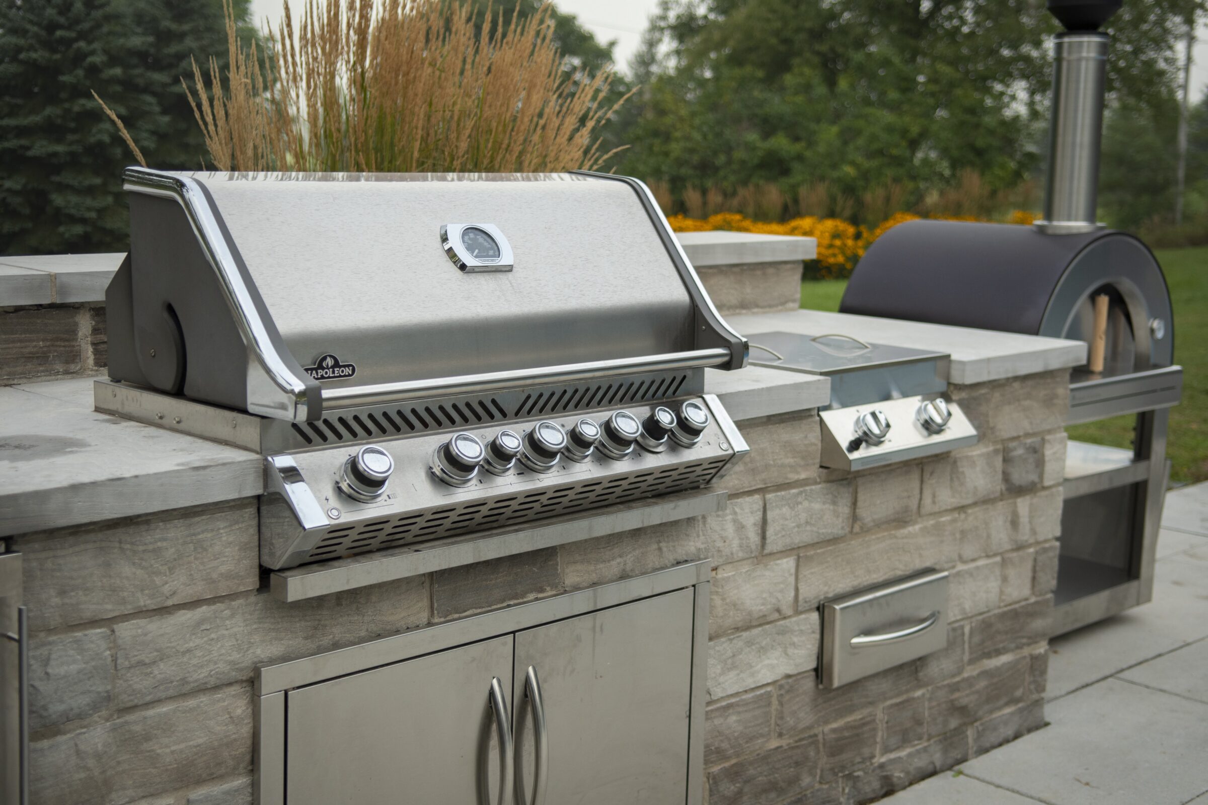 Outdoor kitchen with a stainless steel grill and pizza oven set on a stone countertop, surrounded by greenery and ornamental grasses.